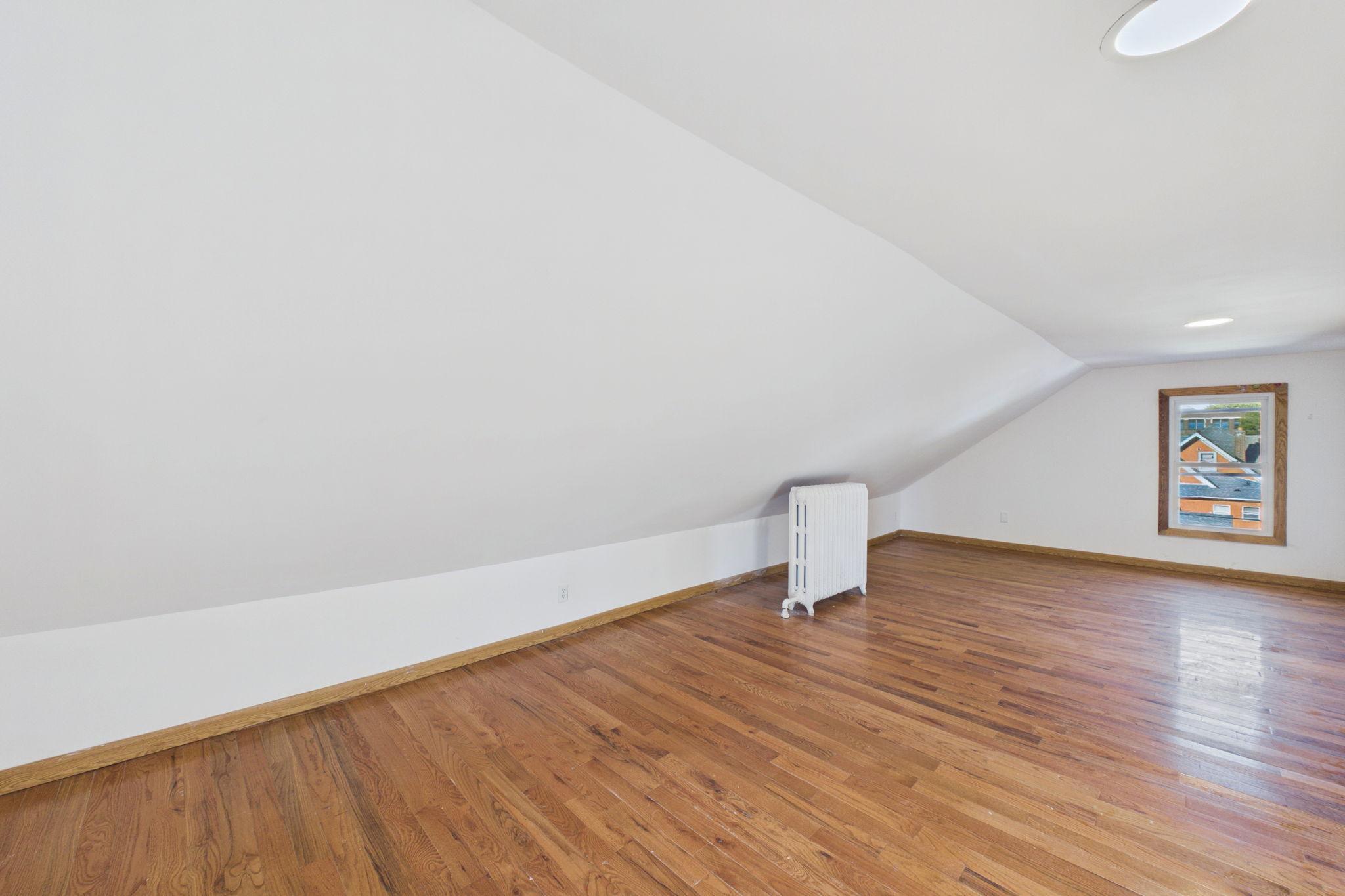 Empty upper-level room with sloped ceilings, hardwood floors, a radiator, and a window.