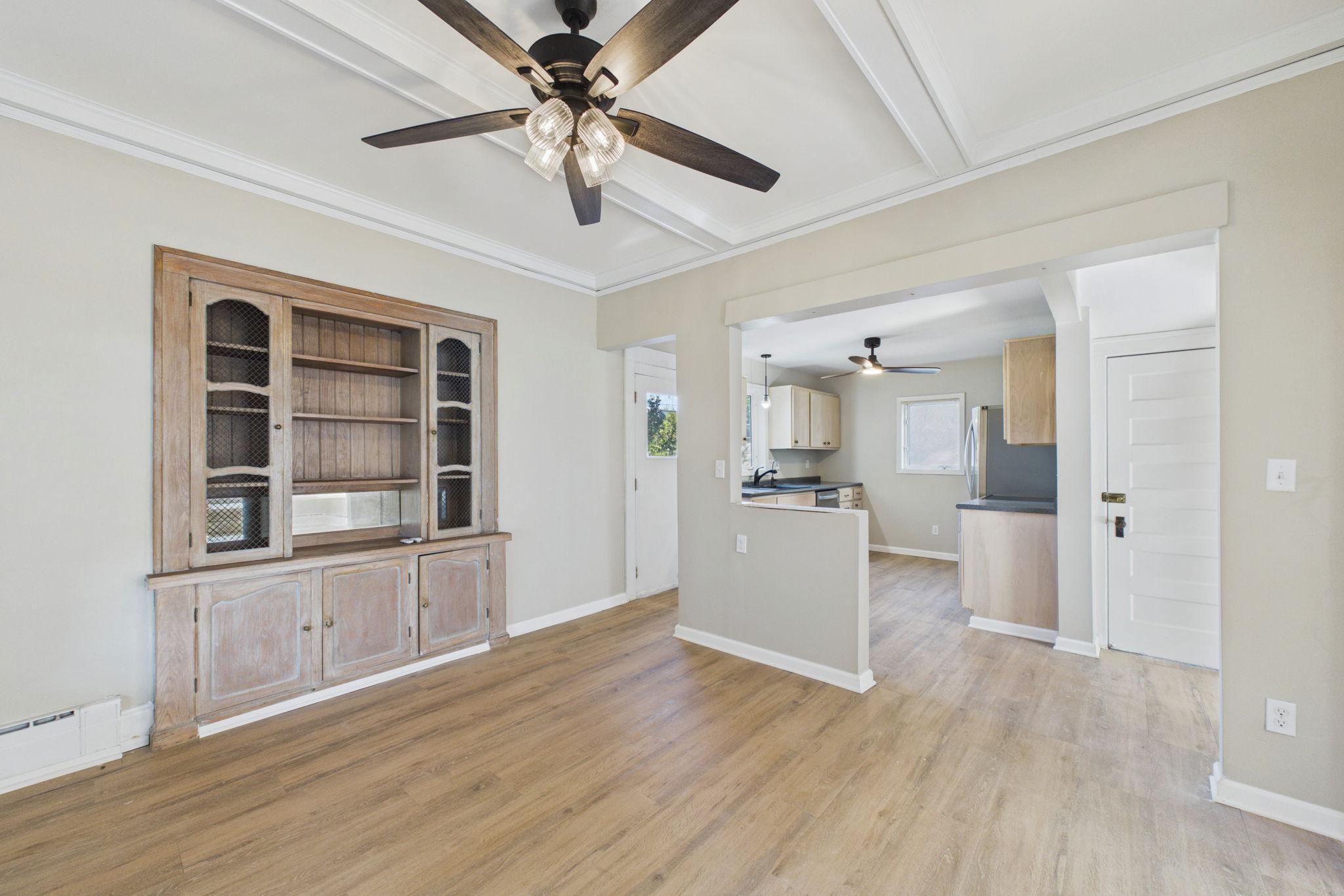 Empty living area with a built-in wooden hutch, coffered ceiling, and open view into the kitchen.