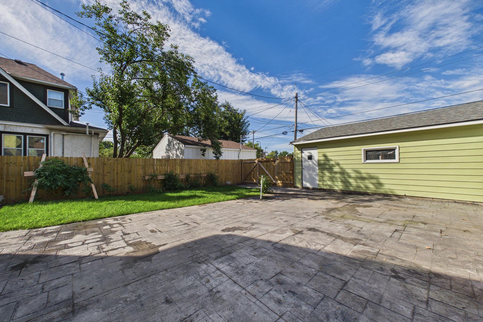 Fenced backyard with a large stamped-concrete patio, lawn, trees, and a light green garage.