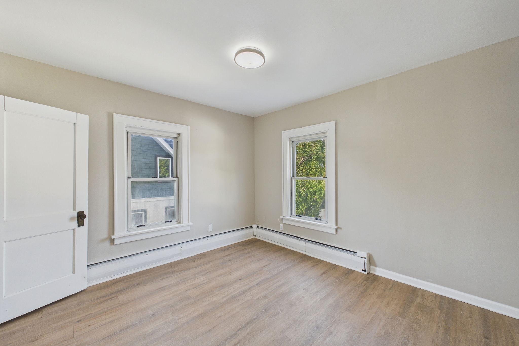 Empty room with light wood-look floors, beige walls, and two windows.