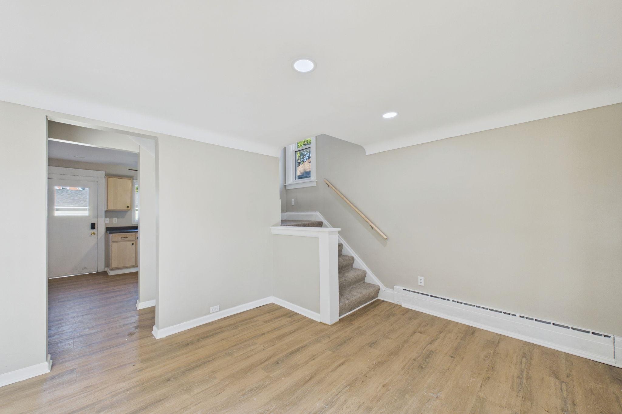 Empty living room with wood flooring and a carpeted staircase beside the entryway.
