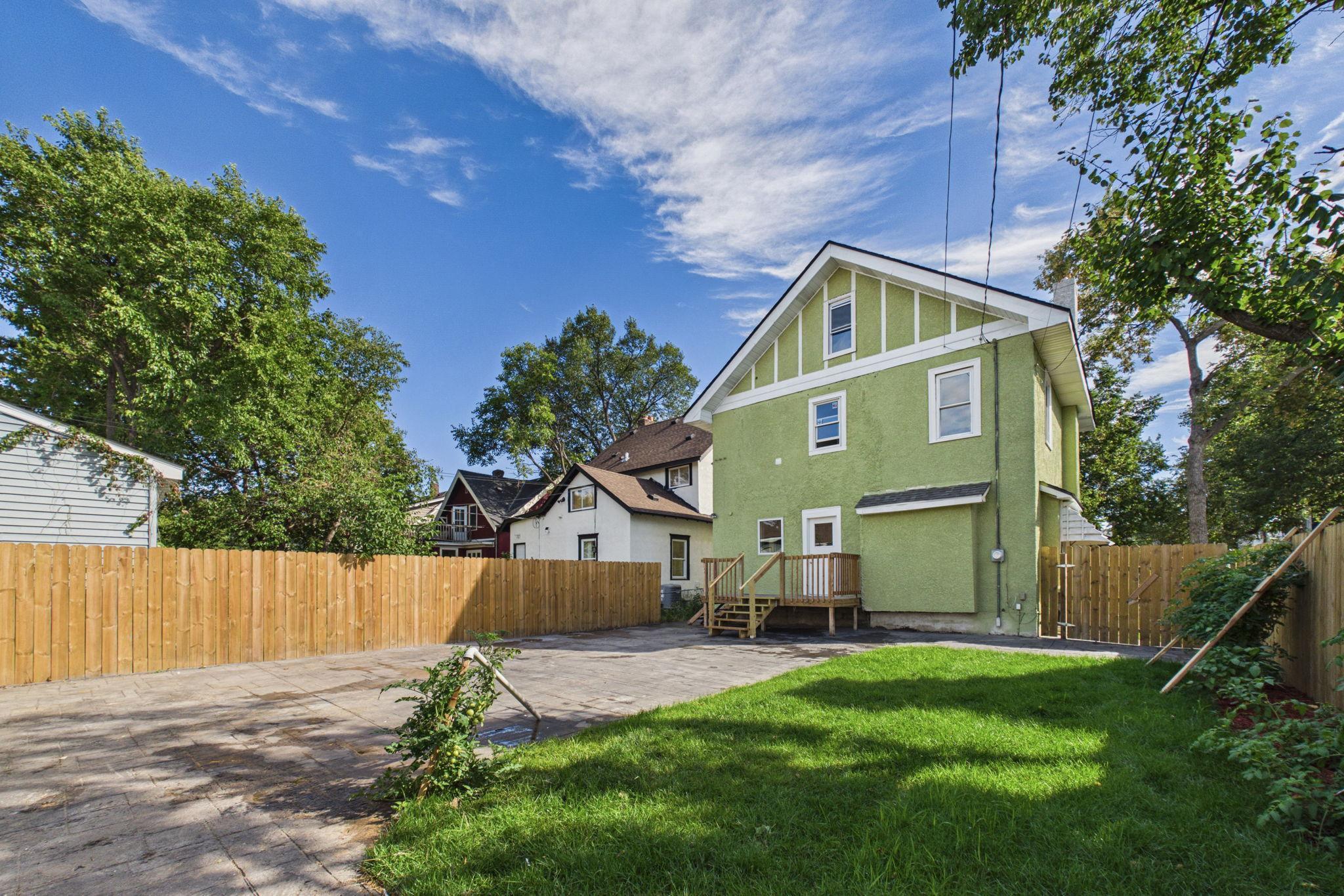 Green two-story house viewed from its fenced backyard, with a wooden deck, paved patio, and lawn.