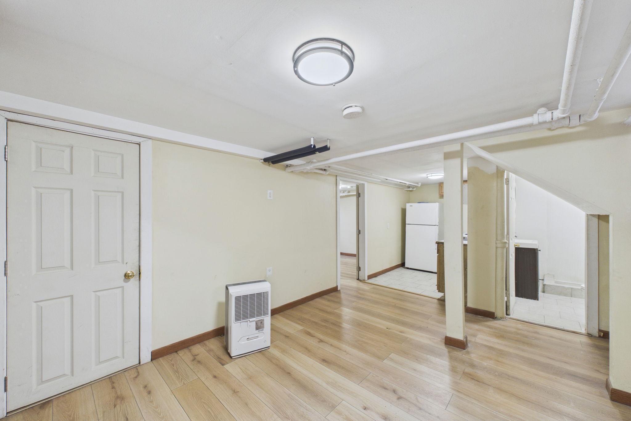 Unfurnished basement room with wood-look flooring, exposed ceiling pipes, and a refrigerator in an adjoining nook.
