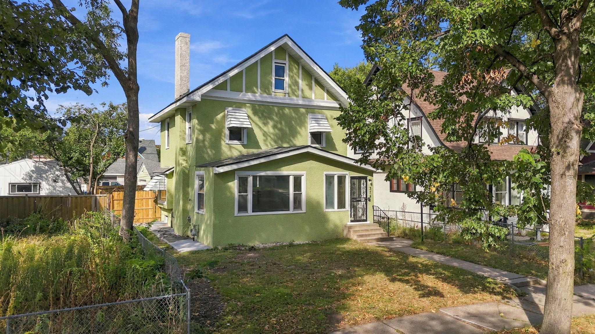 Green two-story home with a steep gable, white trim, and mature trees framing the front yard.