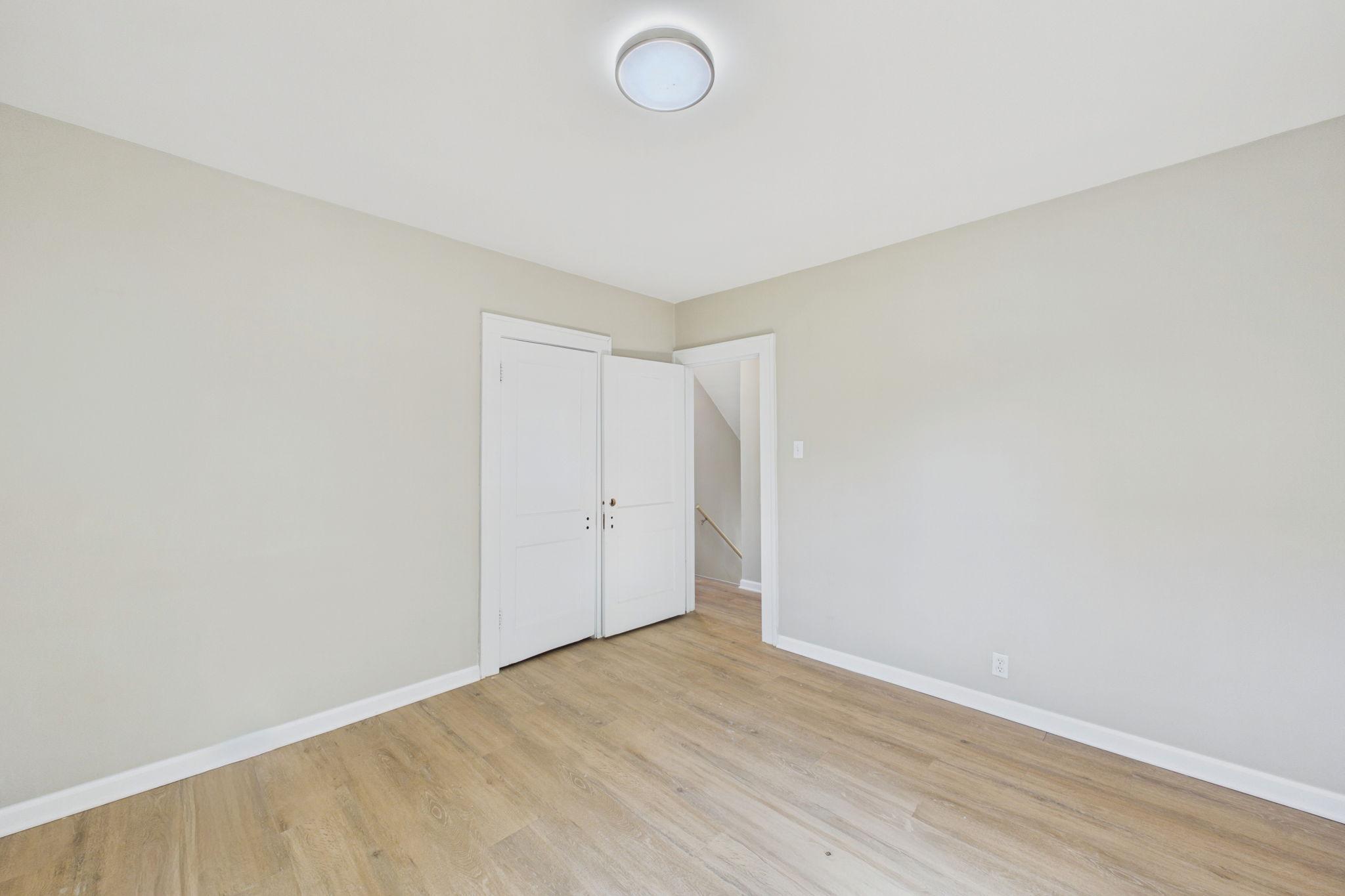 Vacant room with light wood flooring and open doors to a hallway and stairs.