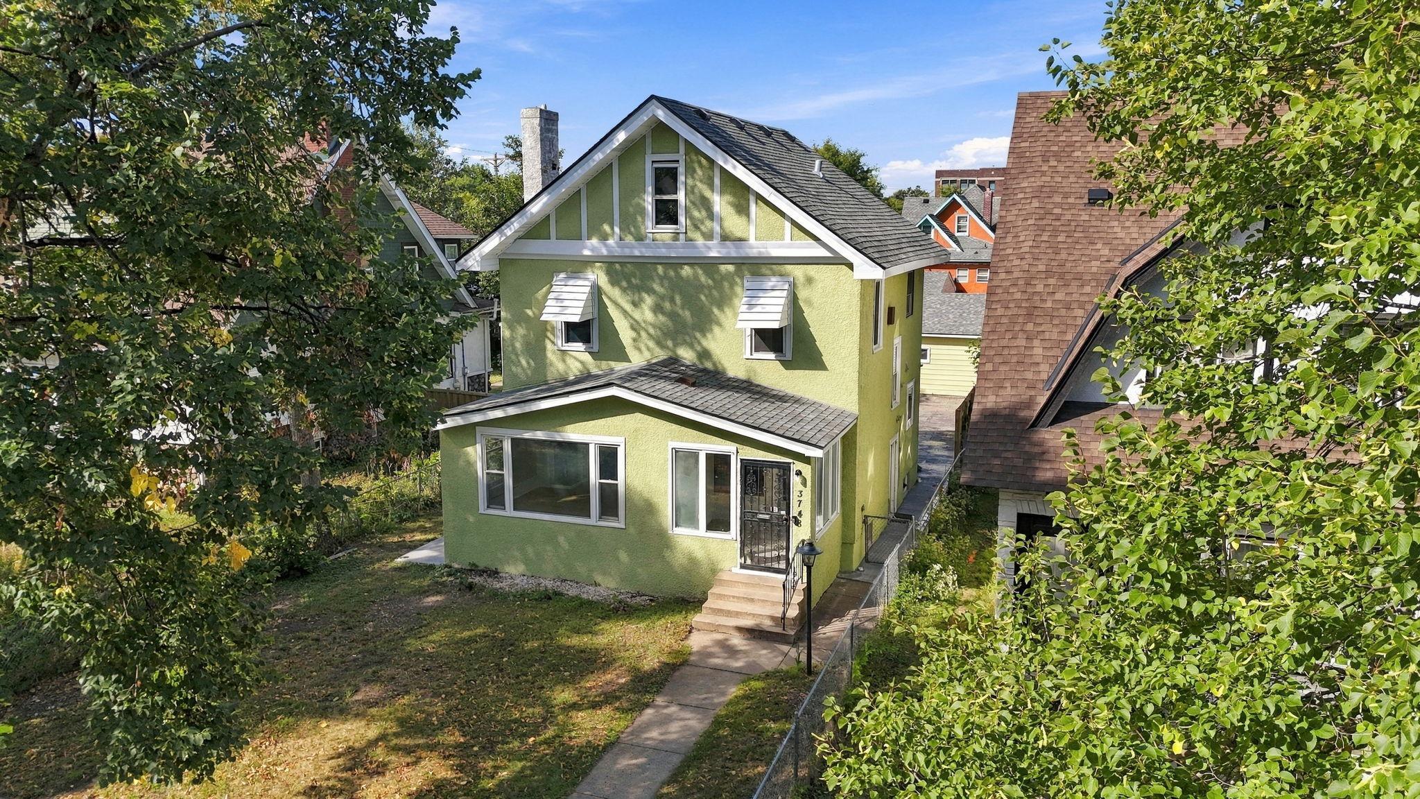 Green two-story house with white trim and steep gabled roof, viewed from above among leafy trees.