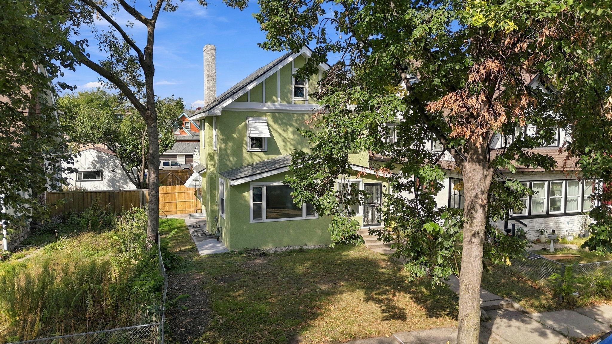 Green gable-roofed house viewed through mature trees, with a fenced yard.