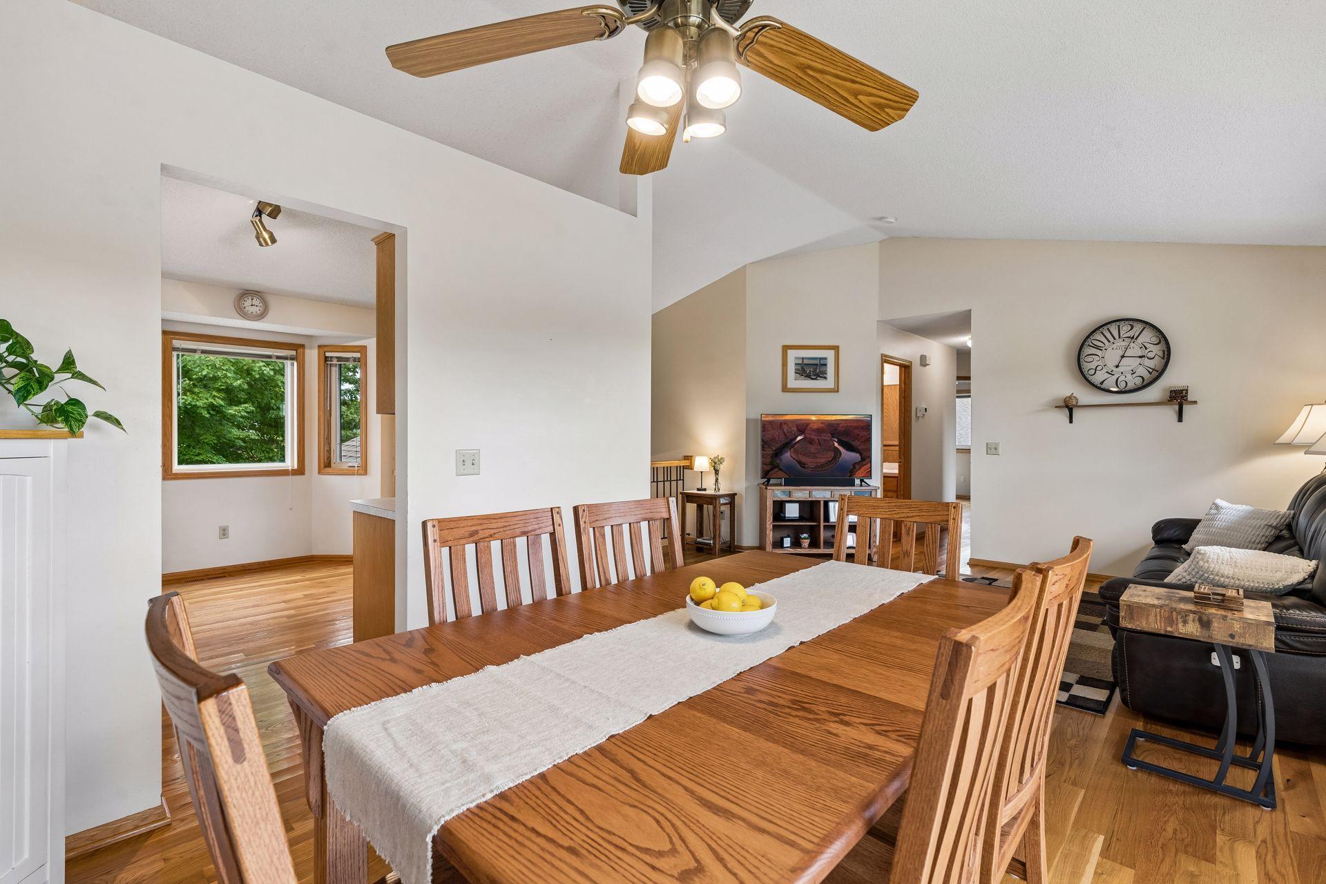 Dining room with a large wooden table and vaulted ceiling, open to a living area.