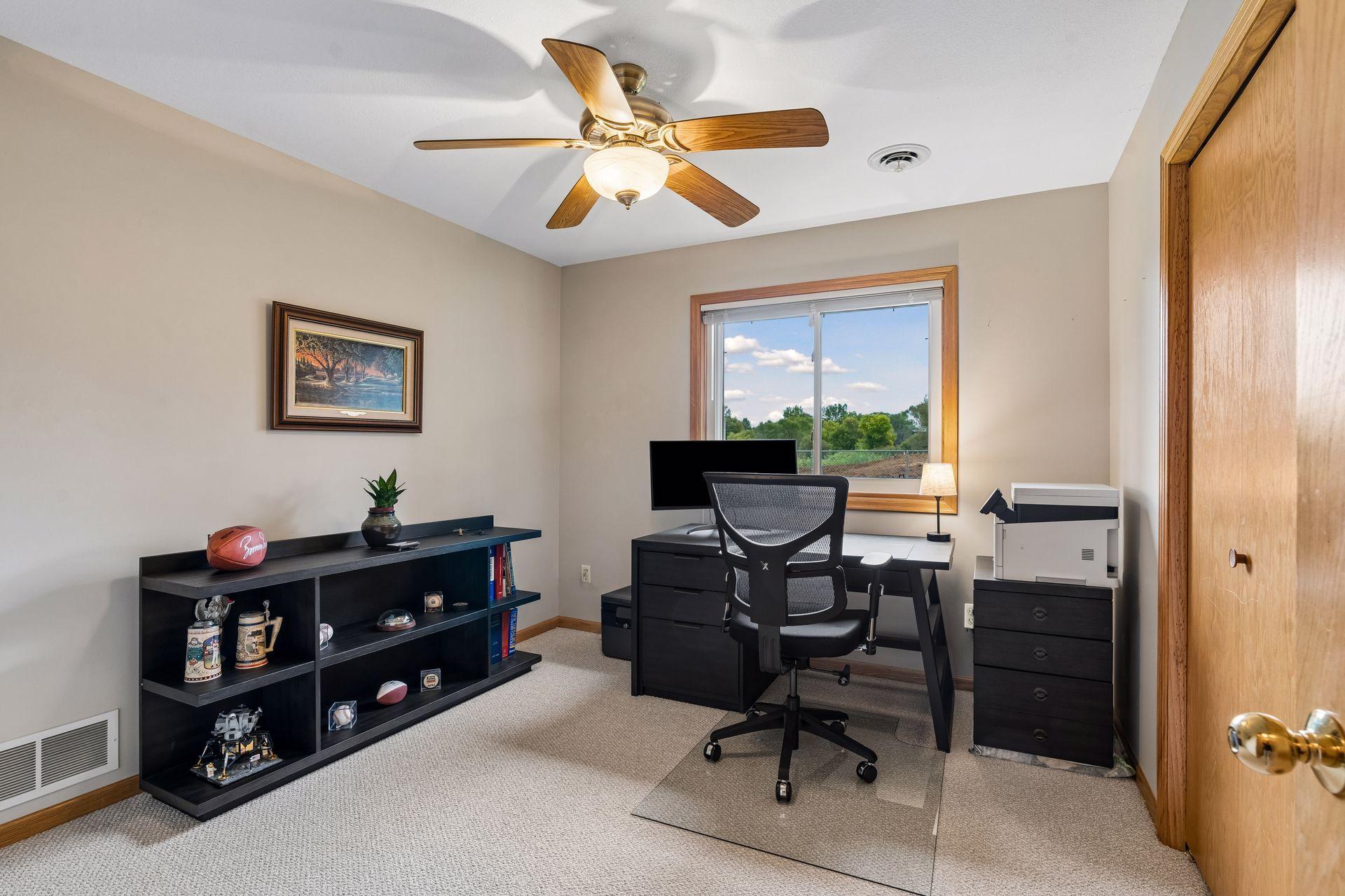 Furnished home office with a black desk, shelving, and a window overlooking trees.
