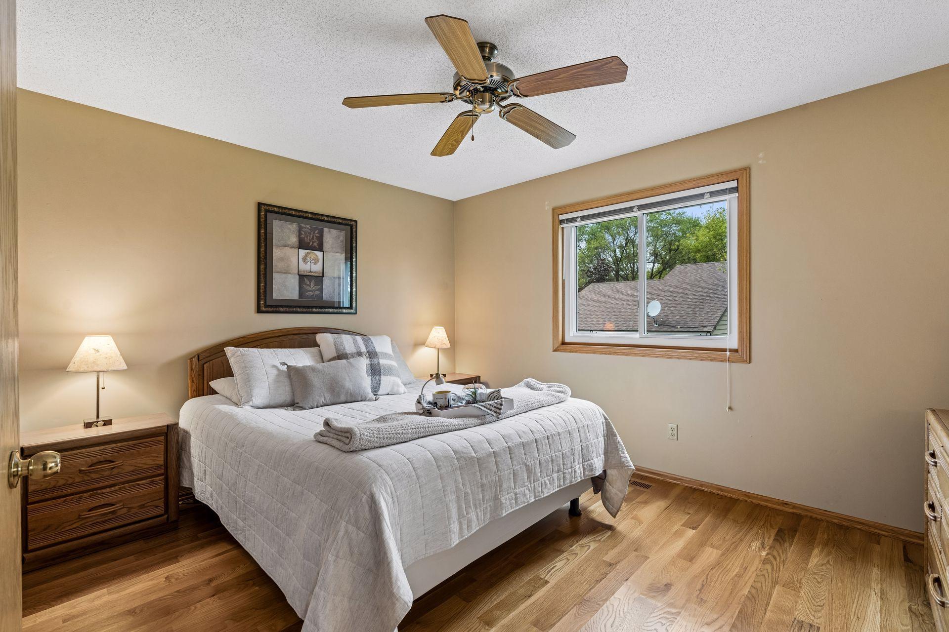 Furnished bedroom with a large white-and-gray bed, hardwood floors, and a wood ceiling fan.