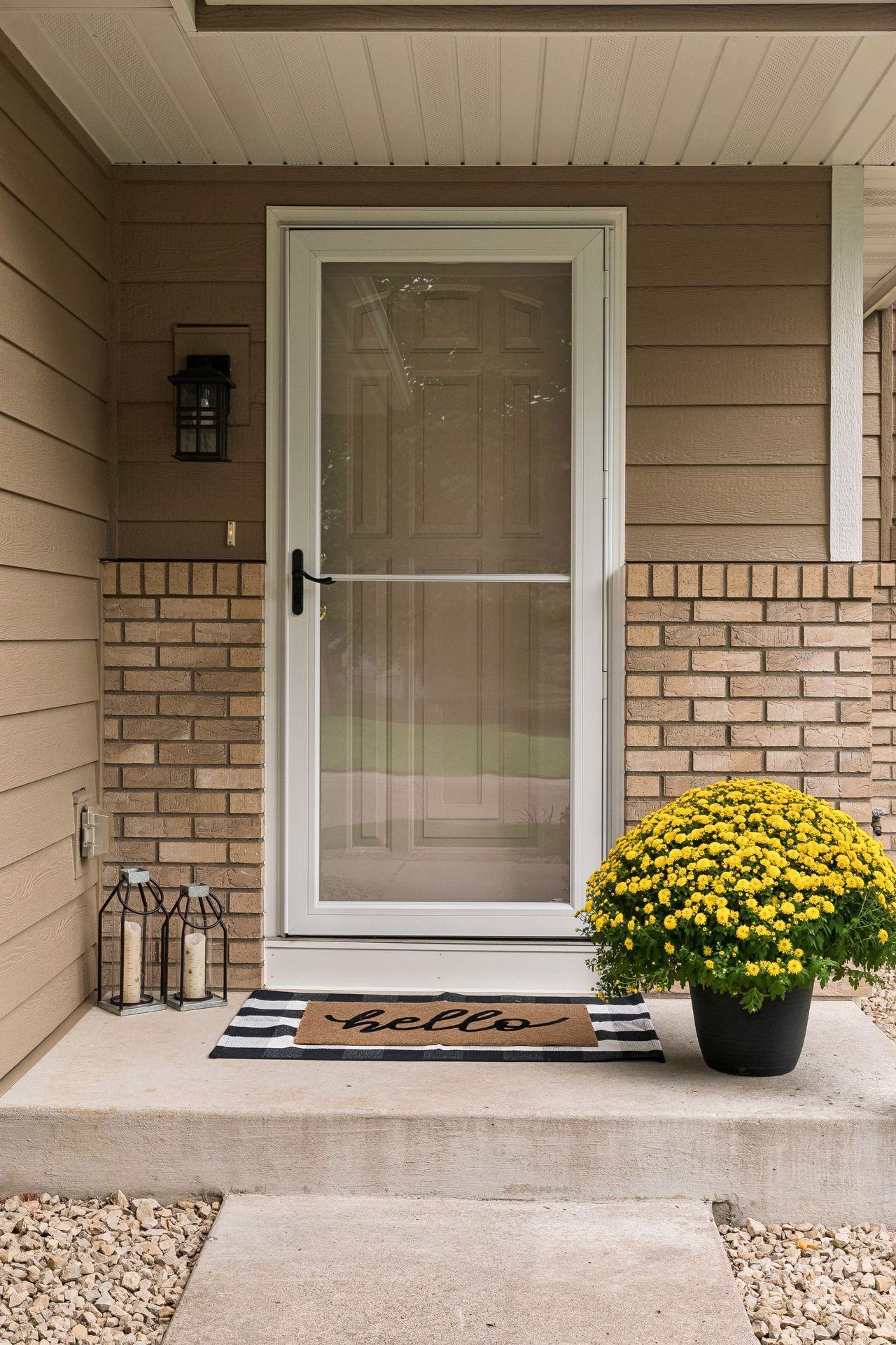Covered front porch with a screen door, brick trim, striped welcome mat, and potted yellow flowers.