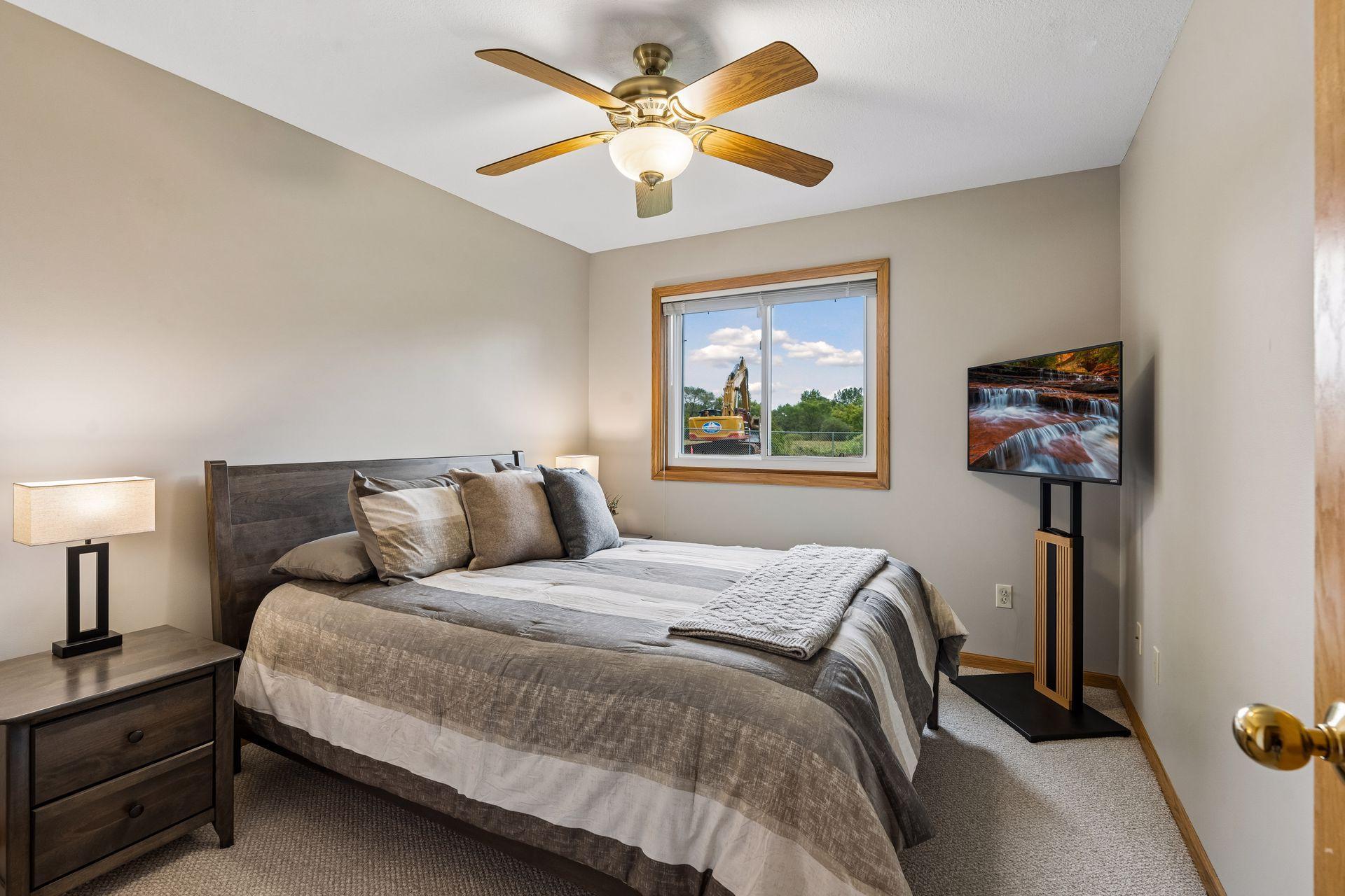 Furnished bedroom with gray bedding, a wood ceiling fan, and an excavator visible through the window.