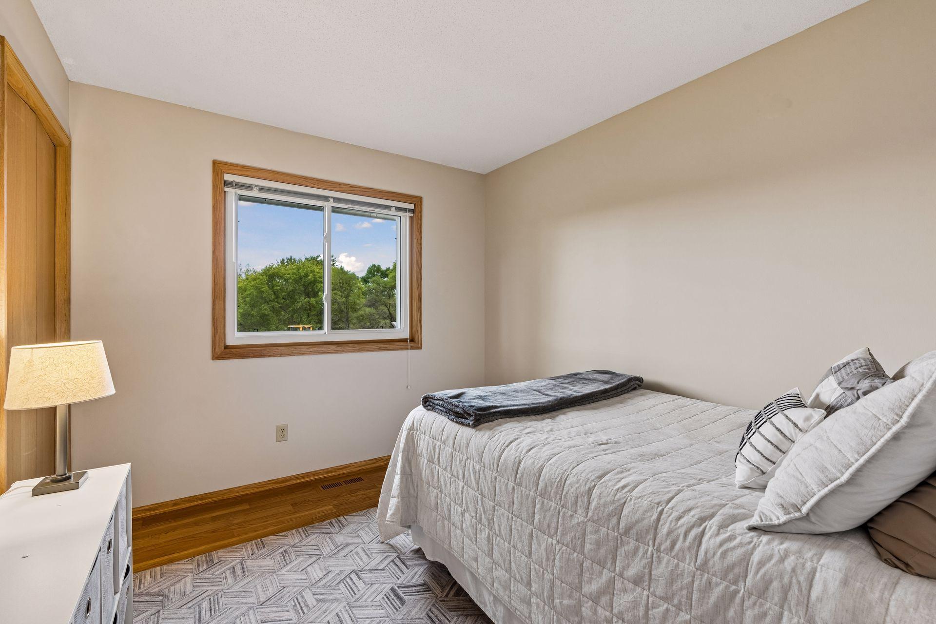 Bedroom with a light quilted bed, wood trim, and a window overlooking trees.