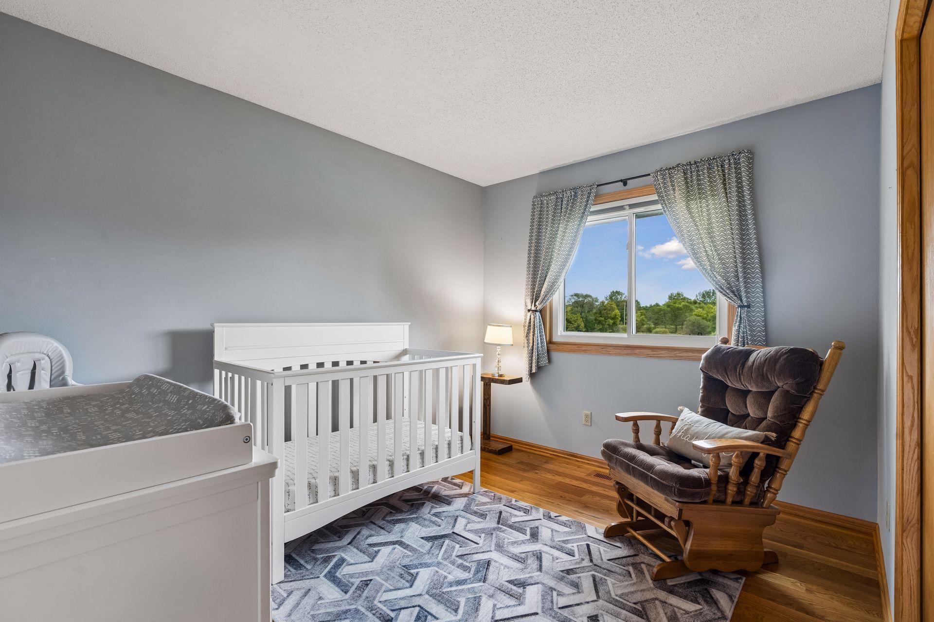 Nursery with a white crib, changing table, wooden rocking chair, and a window overlooking trees.