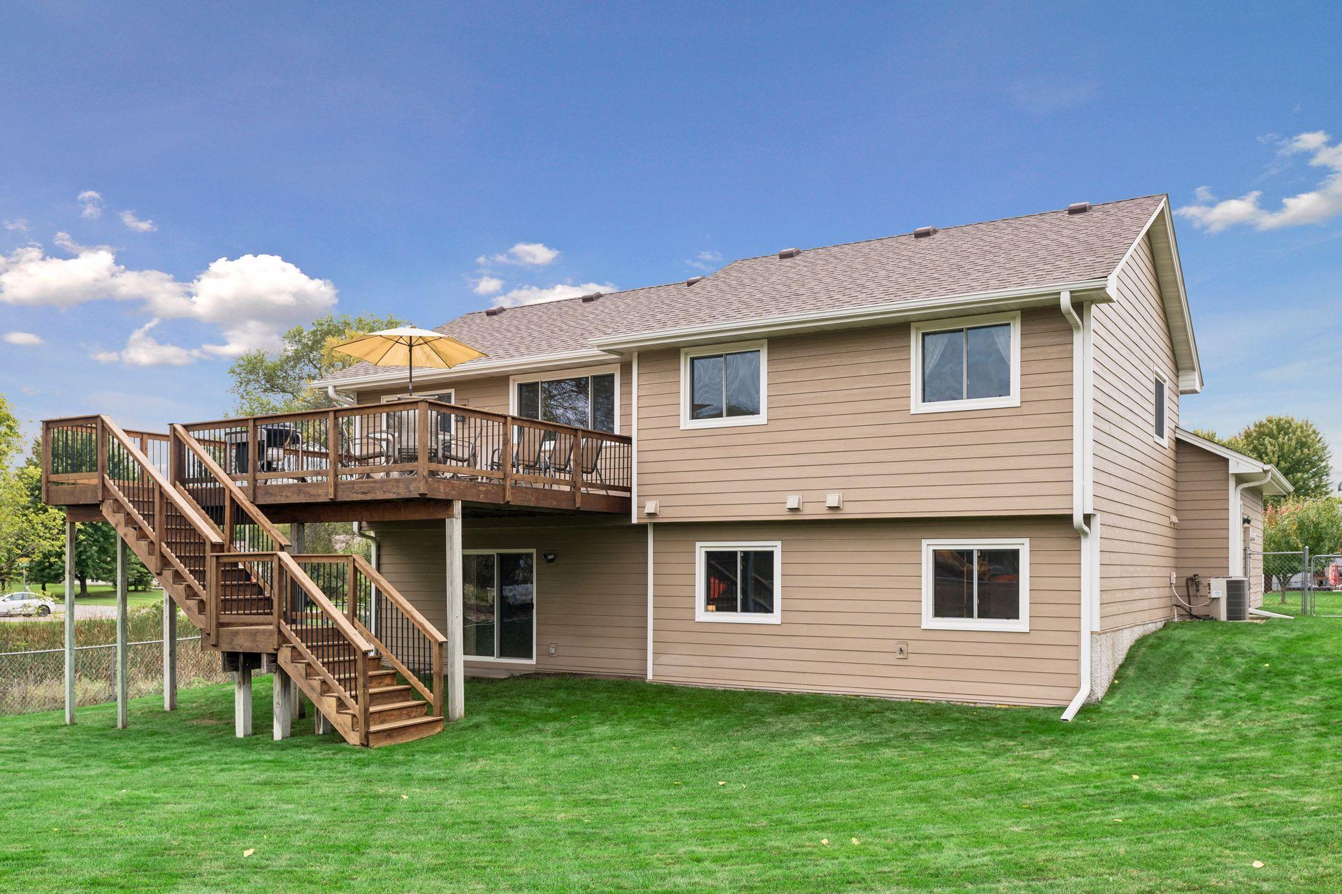Rear of a tan-sided home with a raised deck, wooden stairs, and a patio umbrella.