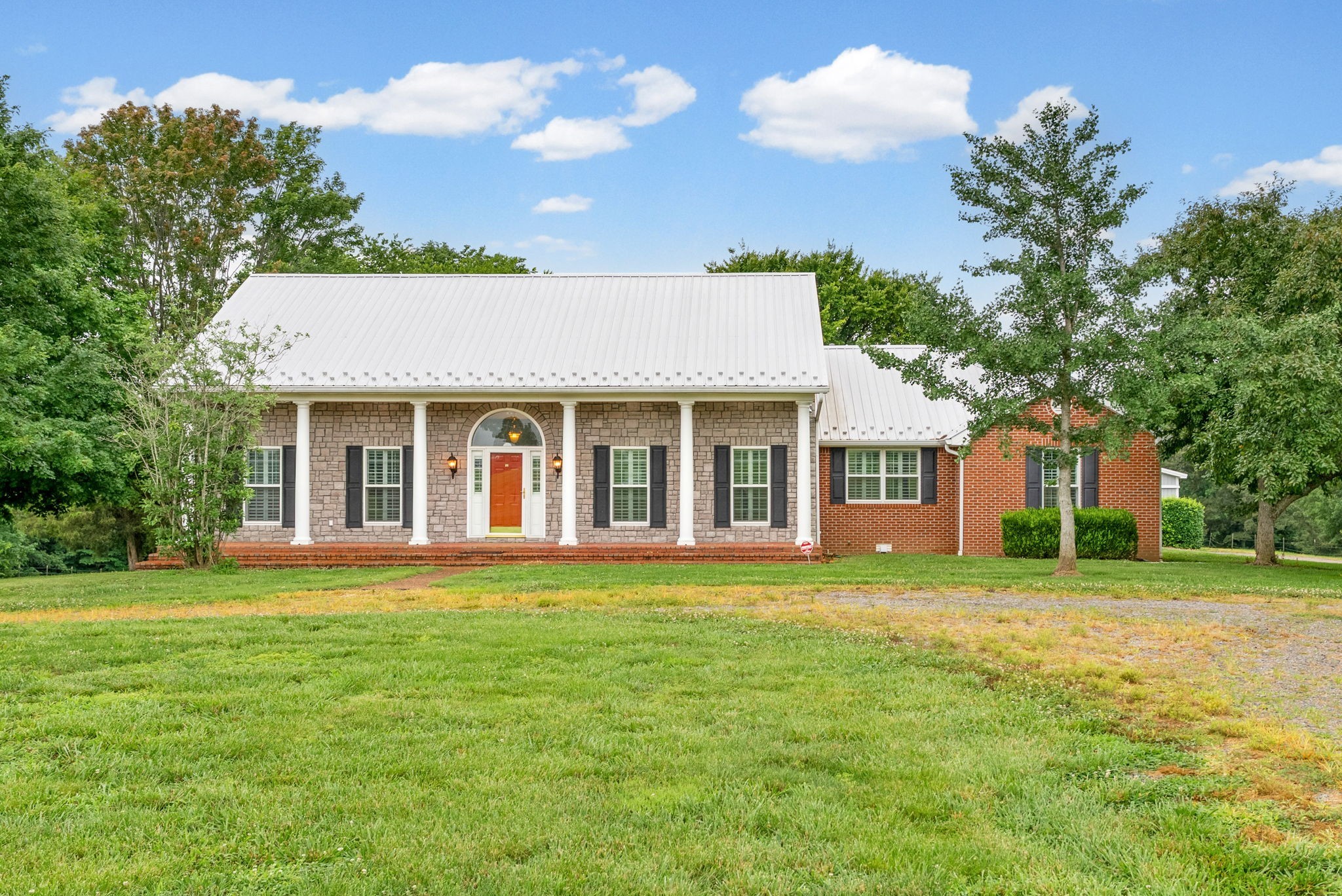 Stone-and-brick house with a white metal roof, columned porch, and broad green lawn.