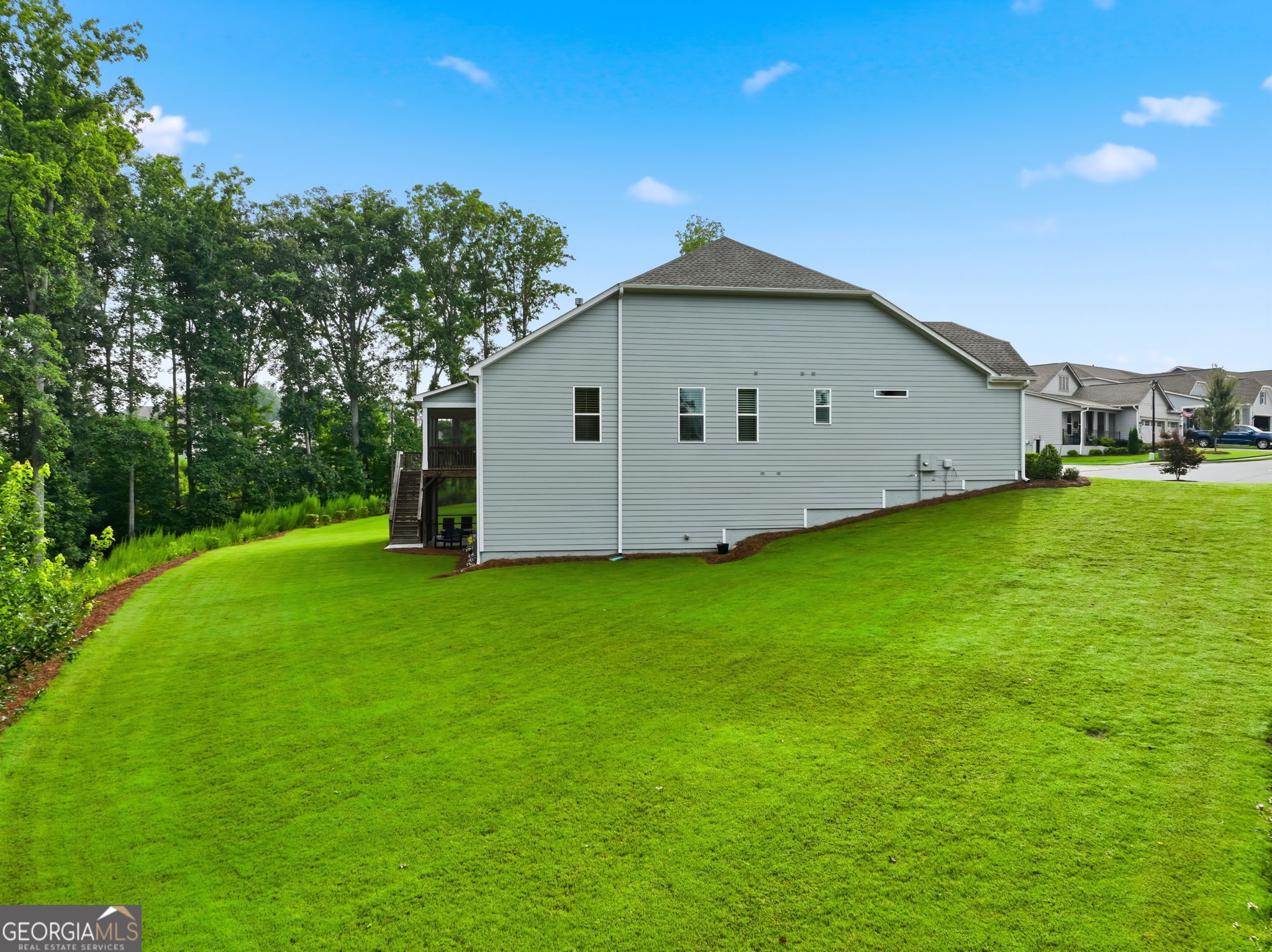 Light-gray house beside a broad sloping lawn, with a raised deck and wooded edge.