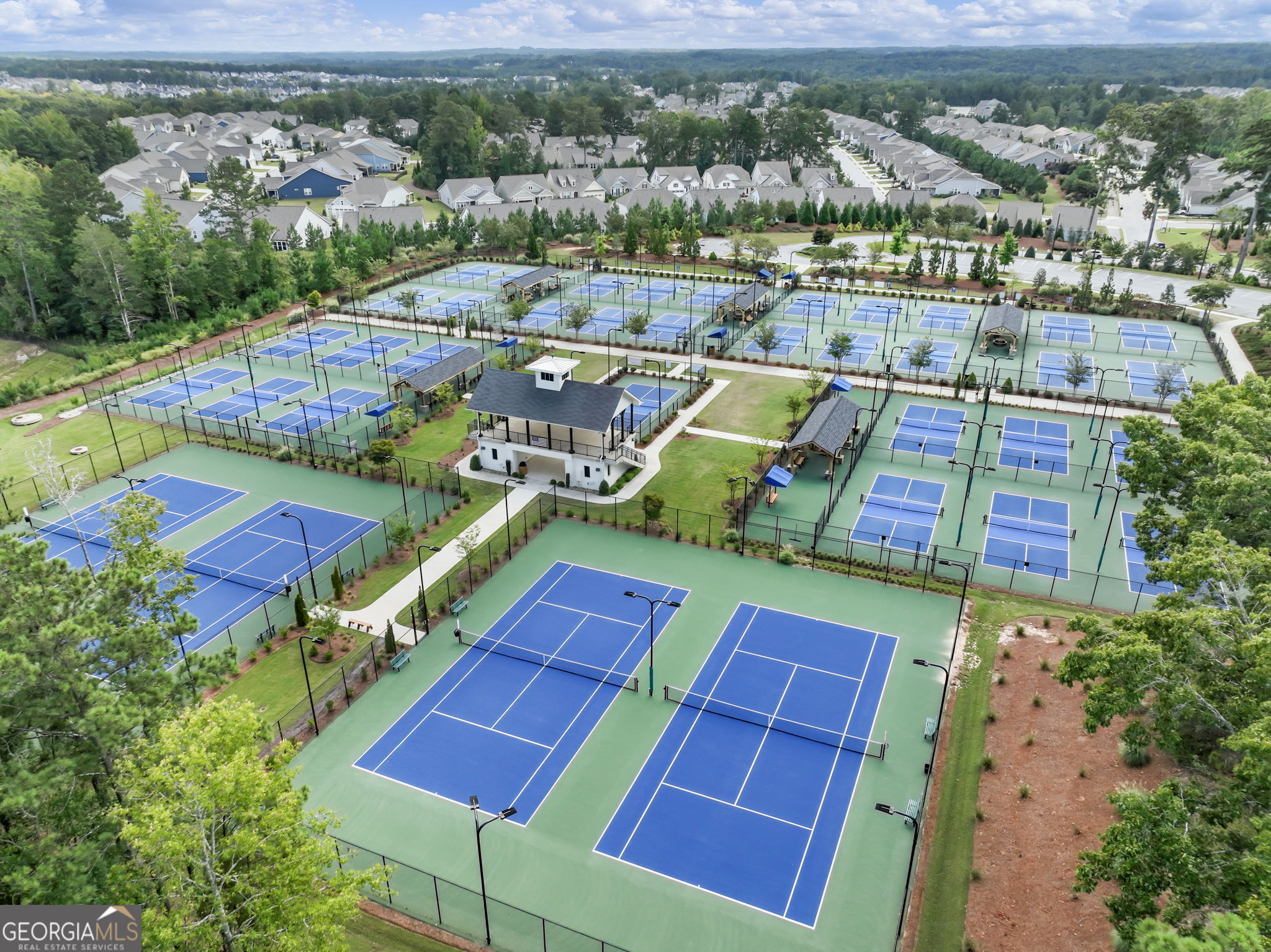 Aerial view of numerous blue pickleball courts beside a central clubhouse and surrounding homes.