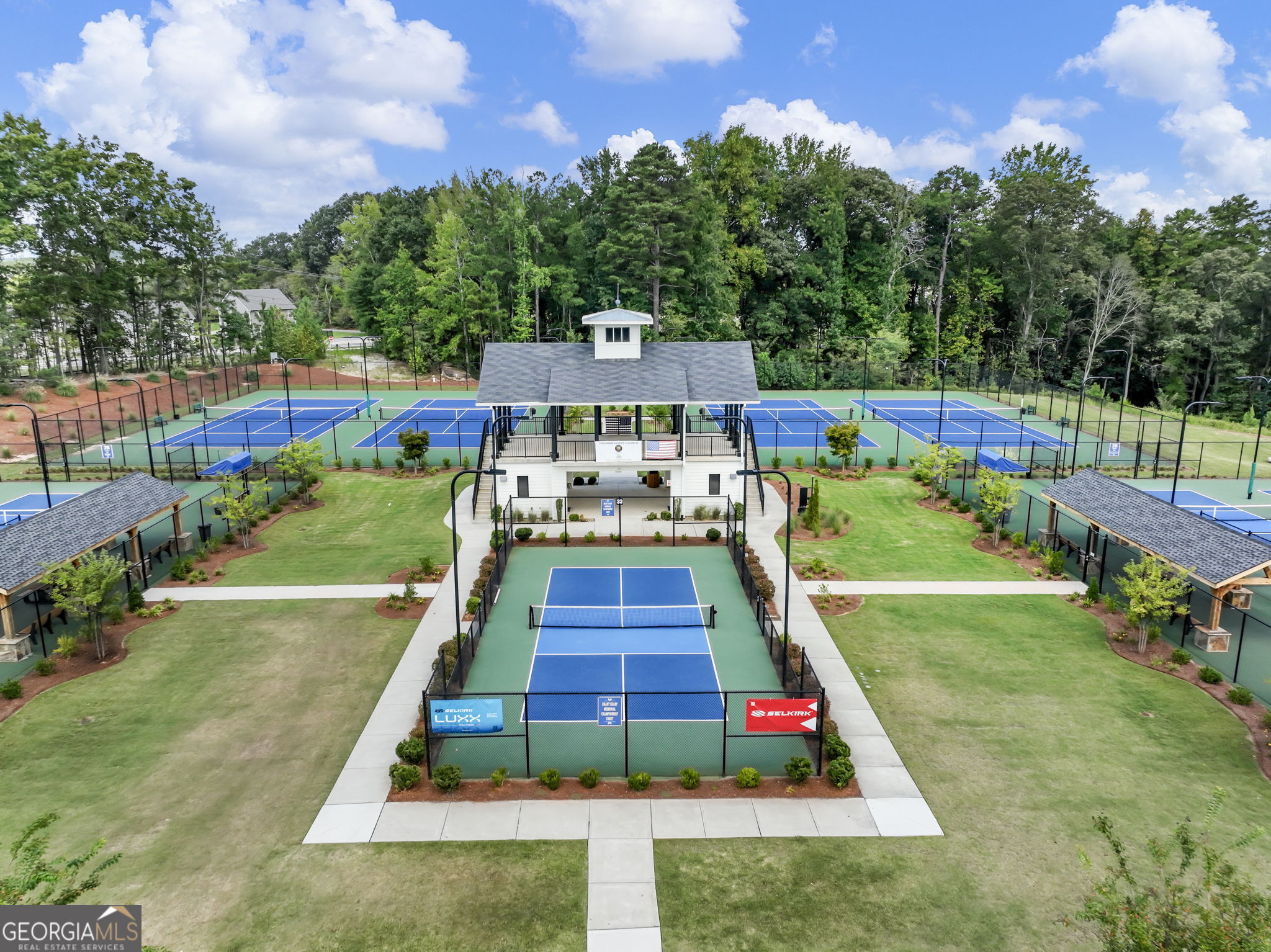 Aerial view of multiple pickleball courts surrounding a clubhouse, bordered by lawns and trees.