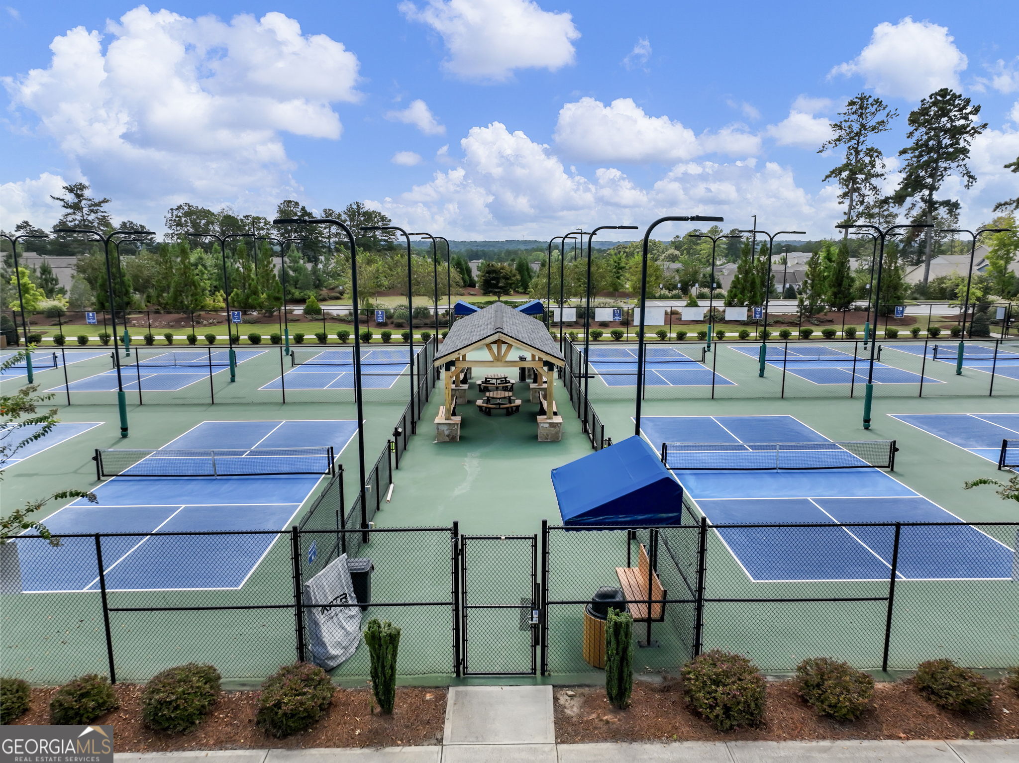 Multiple blue pickleball courts surround a central covered pavilion with picnic tables.