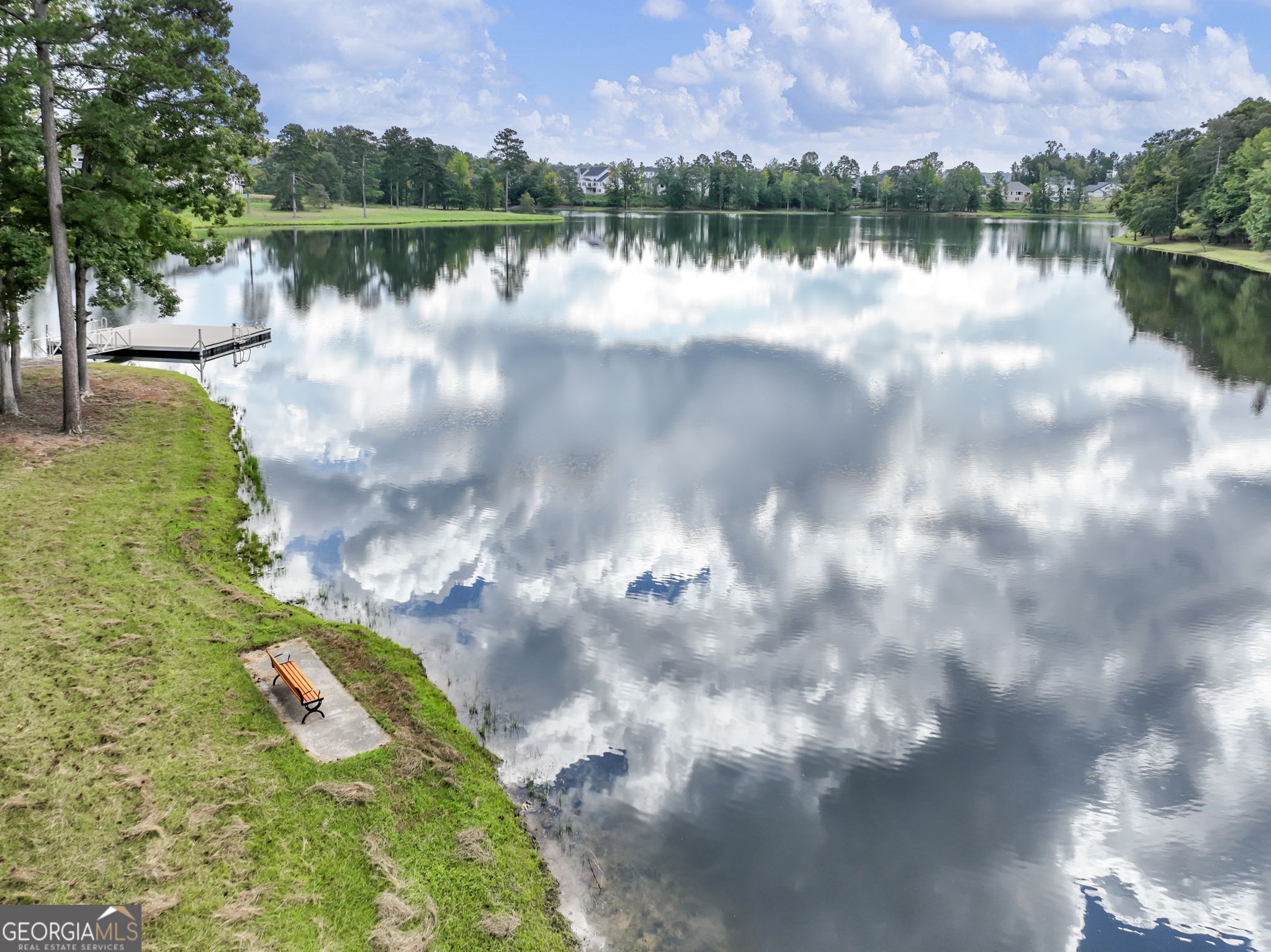 Neighborhood lake reflecting clouds, with a dock and orange bench along the grassy shore.