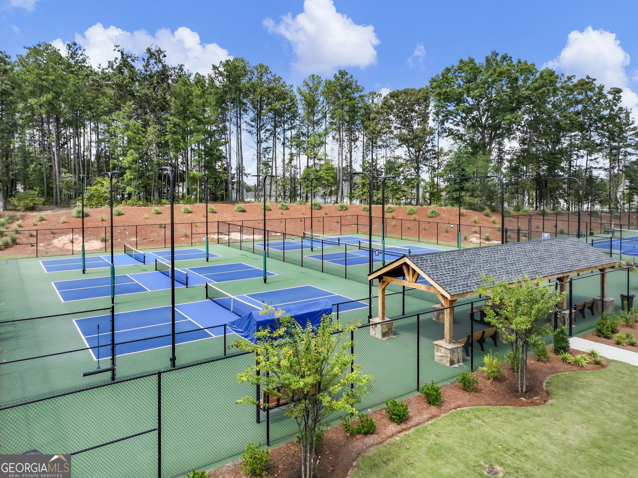 Fenced pickleball courts with a wooden pavilion and wooded landscaping.