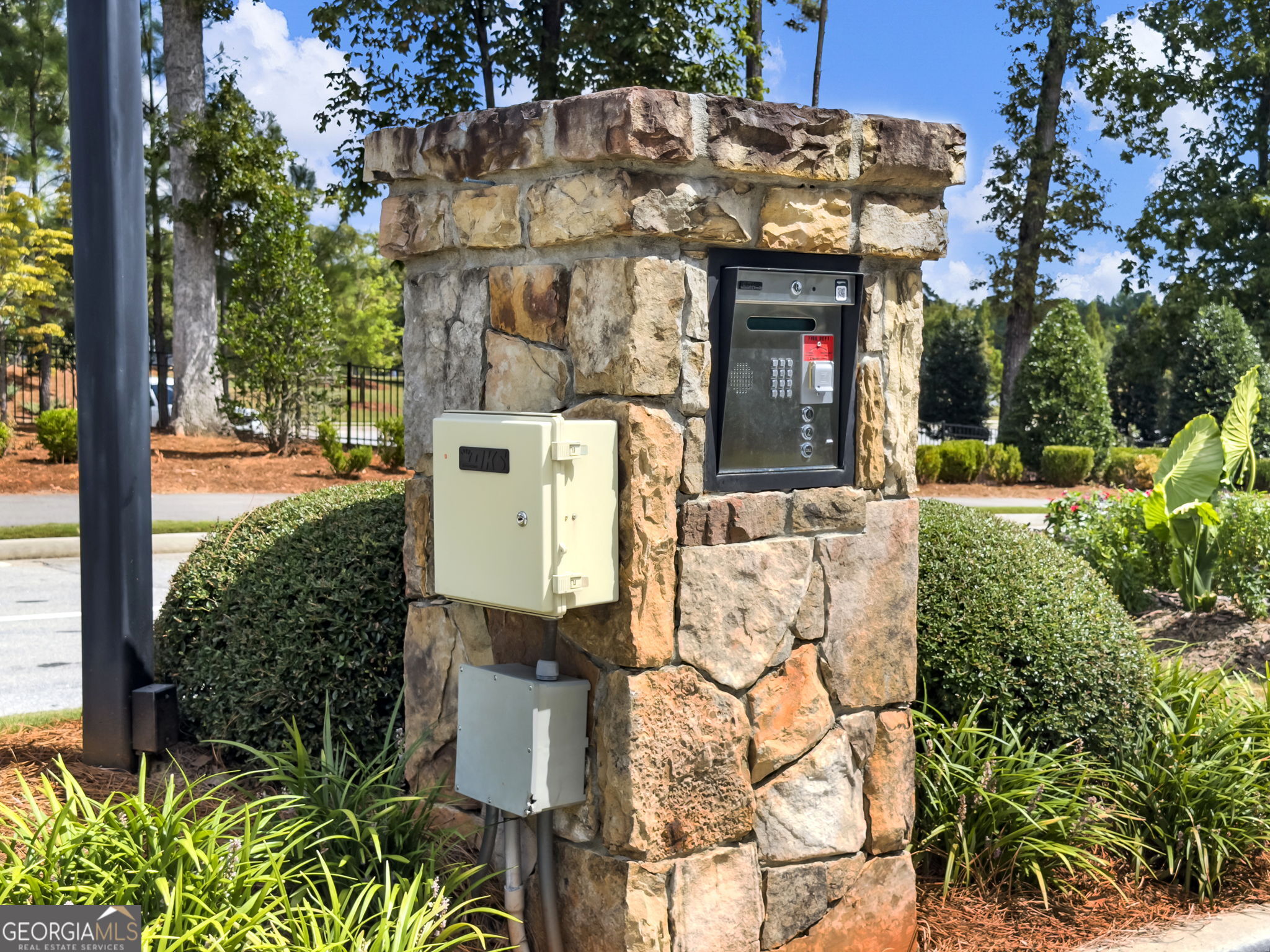 Stone community entrance pillar with a keypad intercom and mailbox, surrounded by landscaped shrubs.