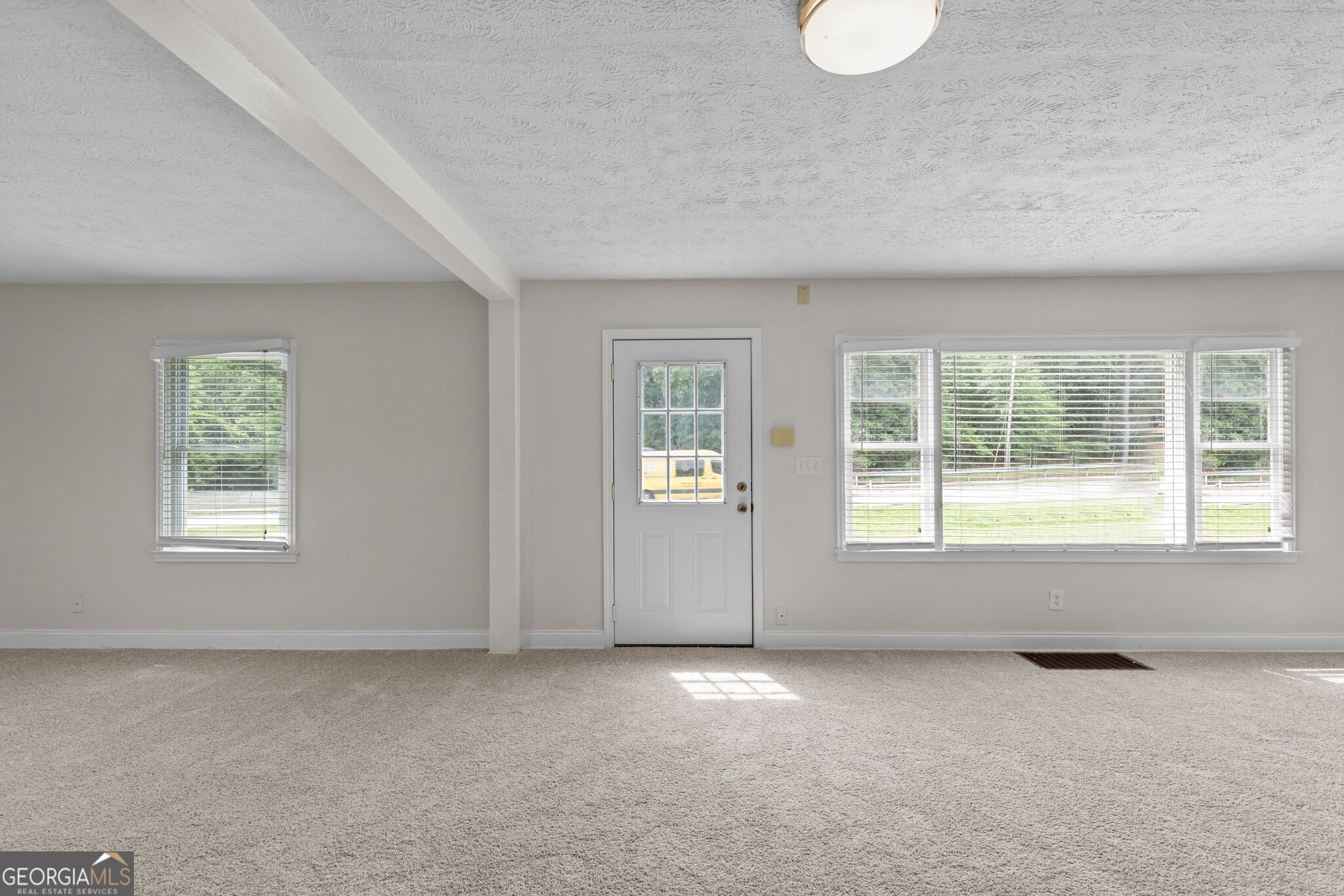 Vacant carpeted room with a glass-paneled exterior door and wide windows with blinds.