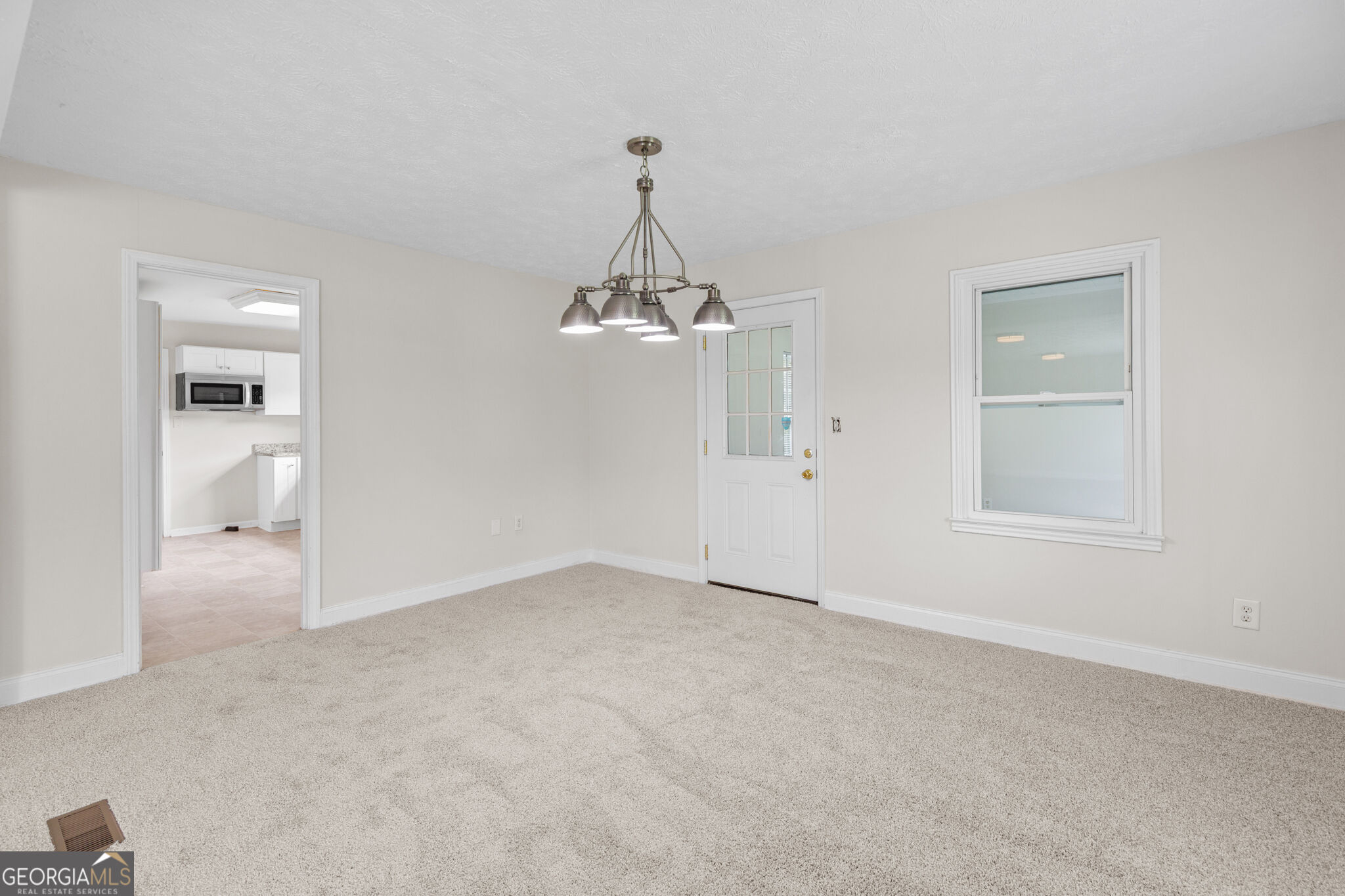 Empty carpeted dining room with a chandelier, window, and glass-panel exterior door beside the kitchen.