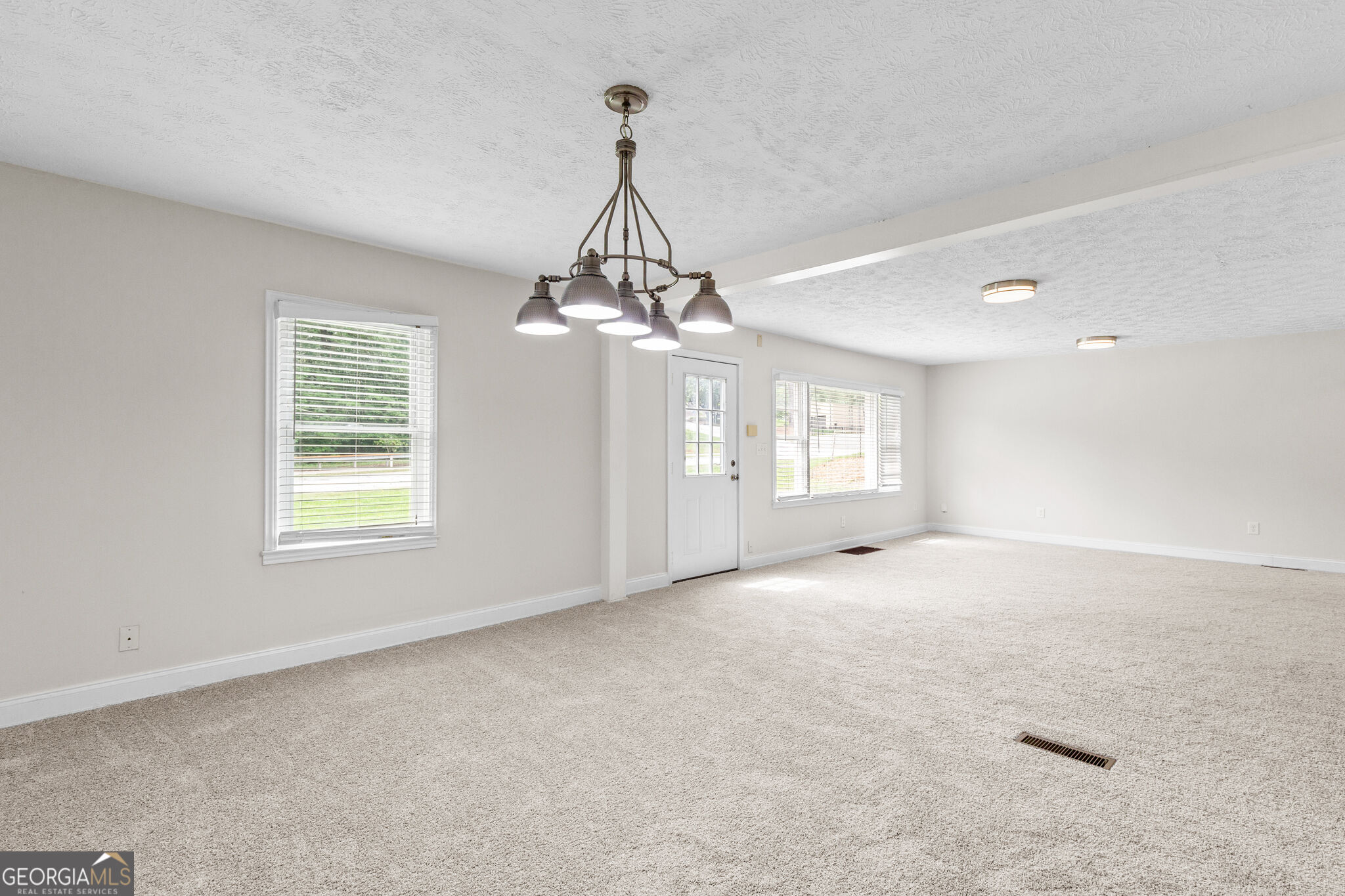 Empty carpeted room with a multi-light chandelier, two windows, and an exterior door.