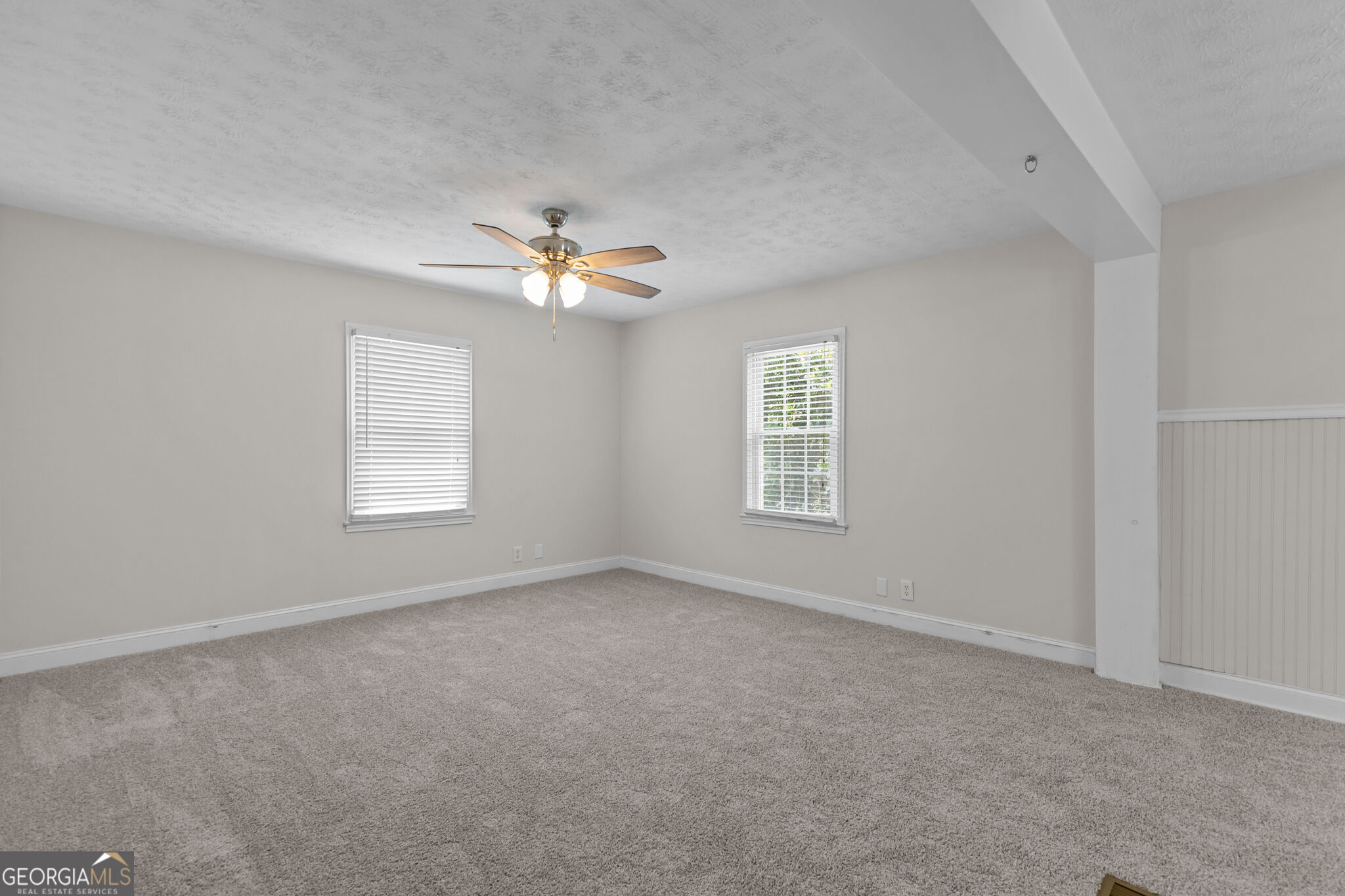 Empty carpeted room with two blind-covered windows, a ceiling fan, and beadboard wainscoting.