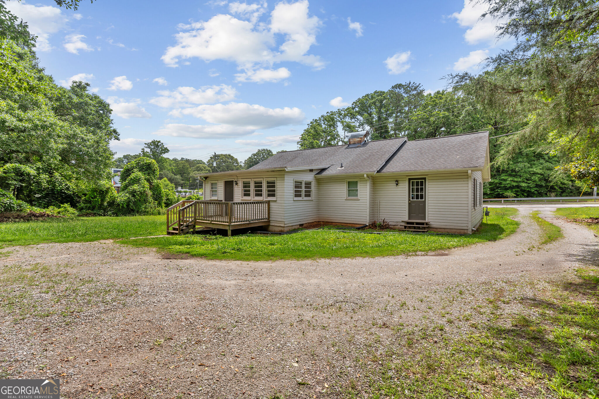Single-story beige house with a small wooden deck, gravel driveway, and wooded surroundings.