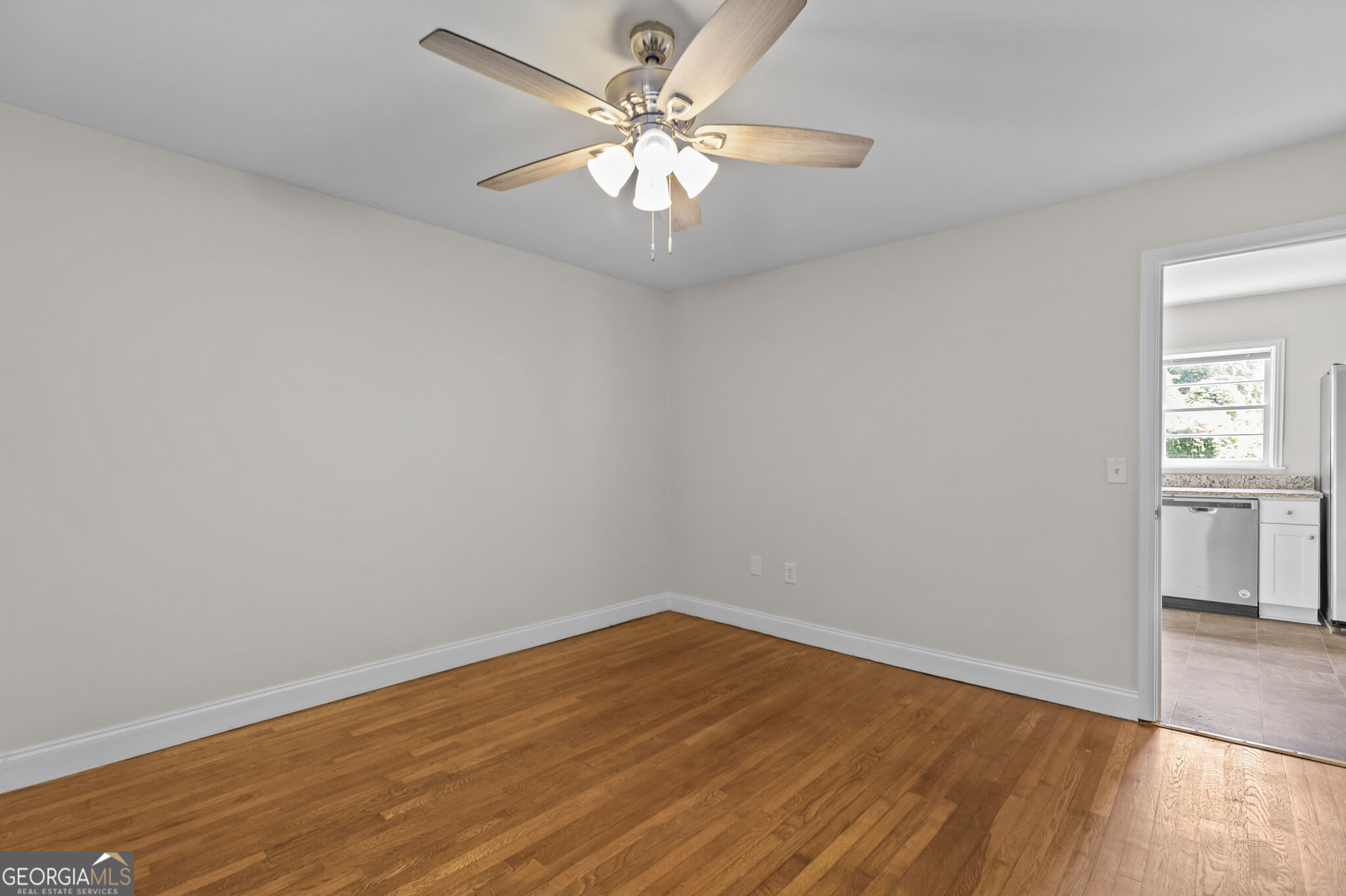 Empty room with hardwood floors and ceiling fan, opening into a kitchen.