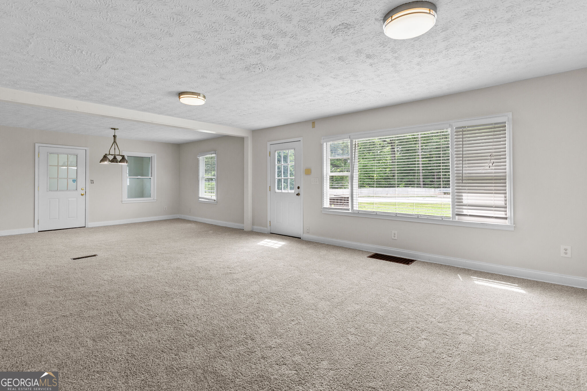 Vacant carpeted living and dining area with two exterior doors and a large window with blinds.