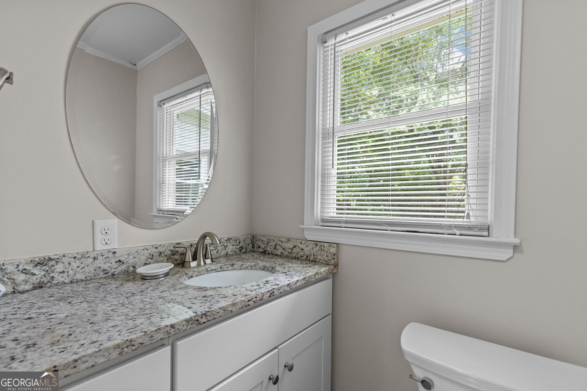 Bathroom vanity with granite countertop, oval mirror, and a bright window beside the toilet.