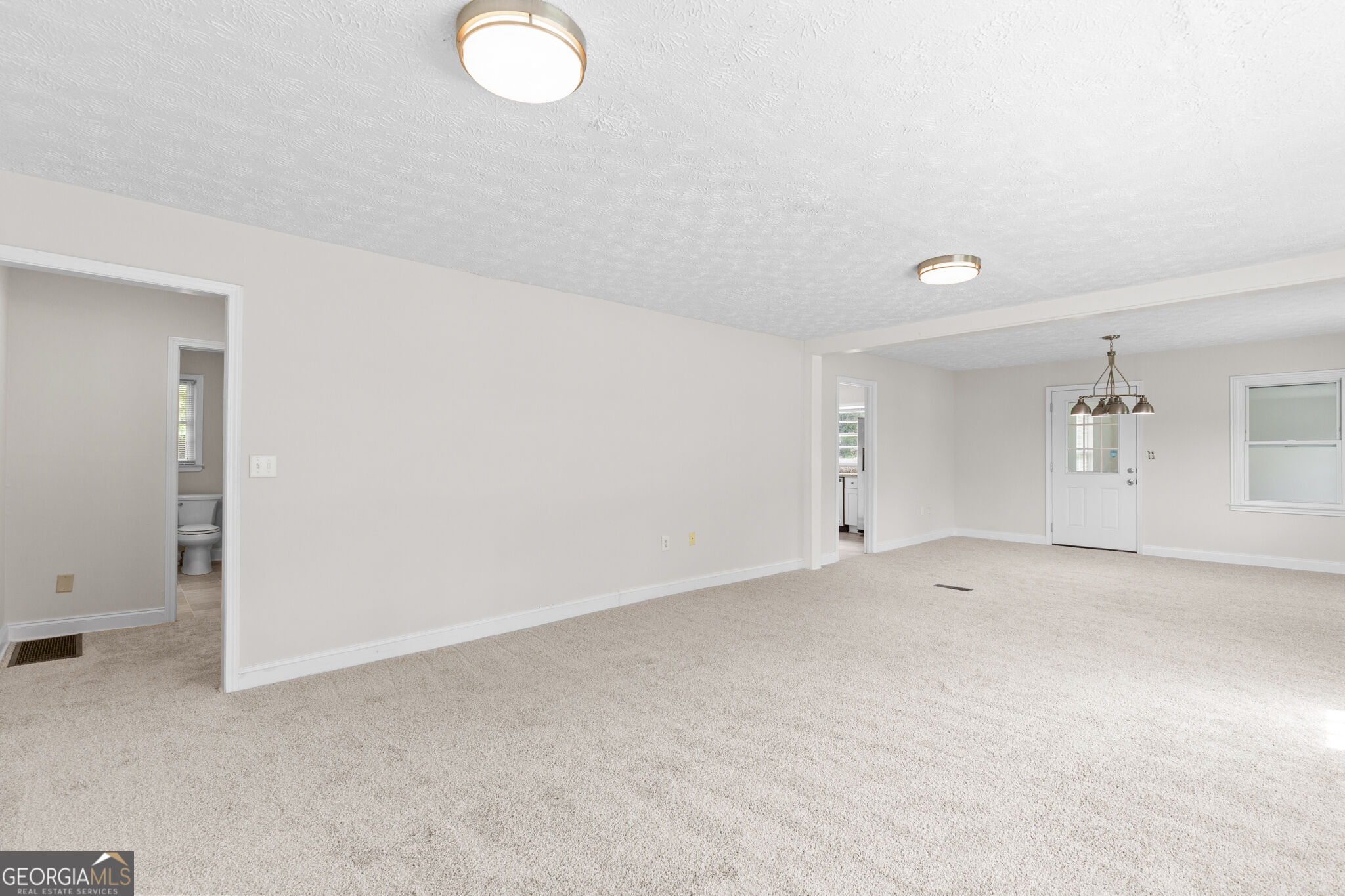 Empty carpeted living and dining area with ceiling lights and a bathroom doorway.