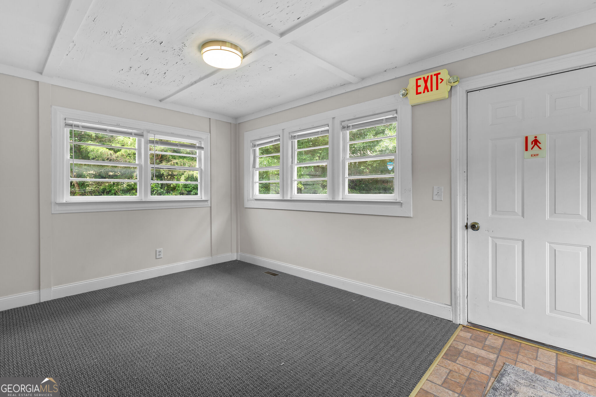 Empty carpeted room with multiple windows and an exit door.