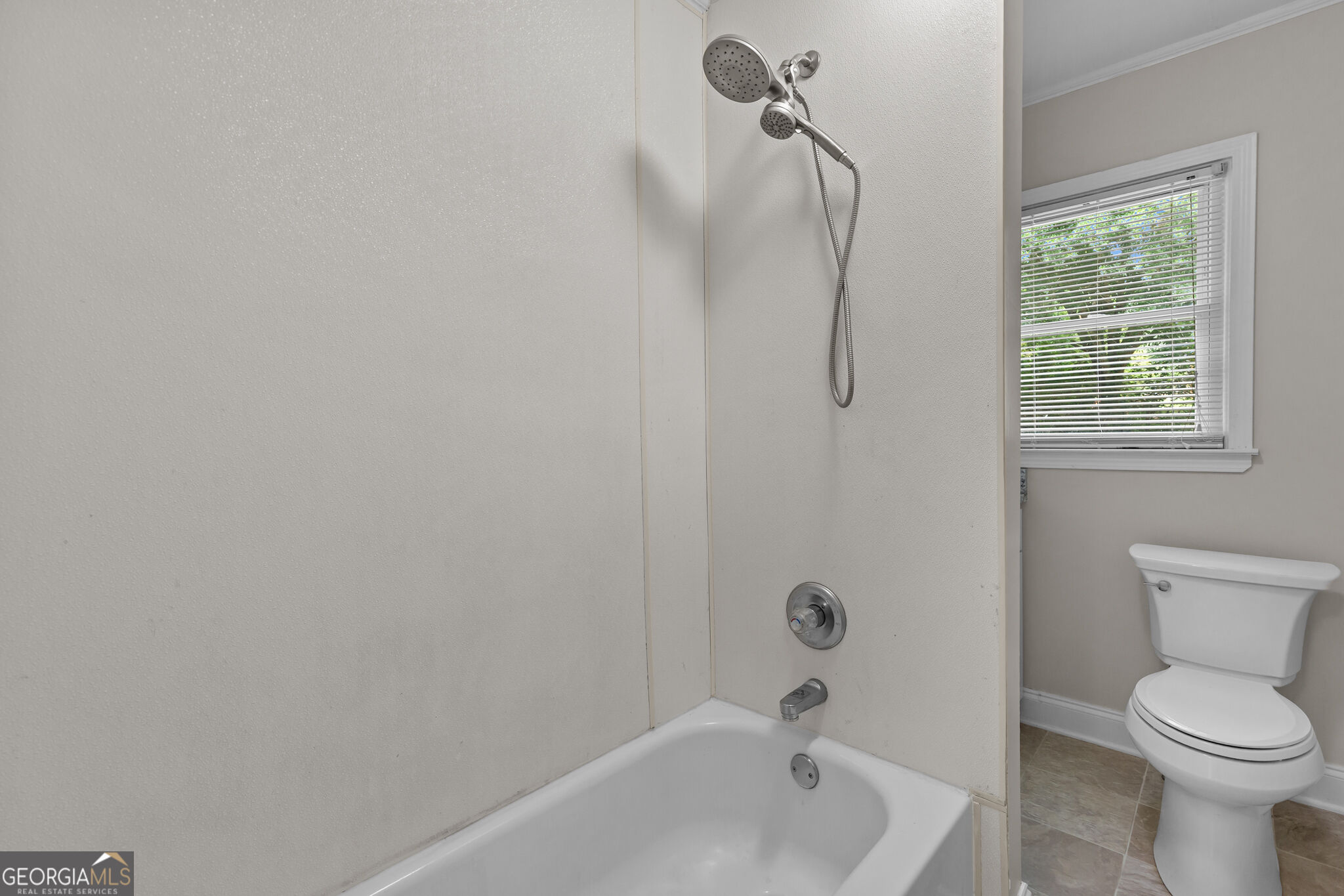 Bathroom with a white bathtub and handheld shower beside a toilet and window with blinds.