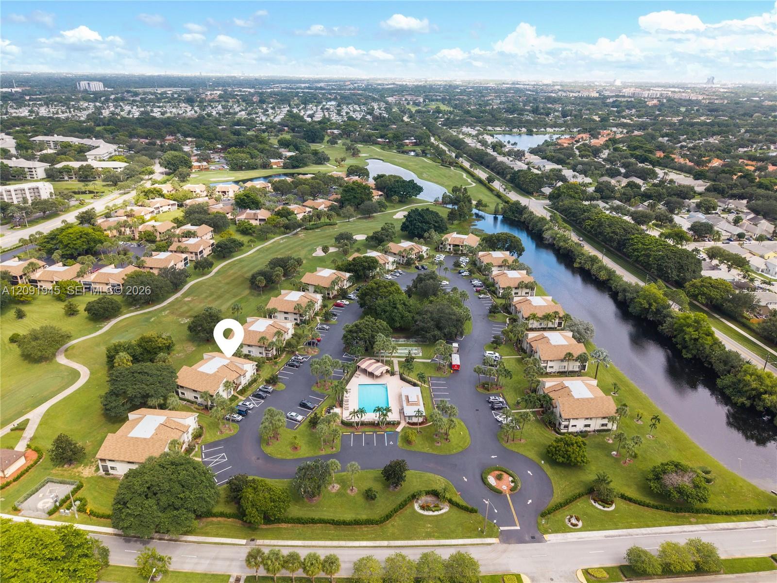 Aerial view of condominium buildings beside a golf course and canal, with a location pin marking one building.