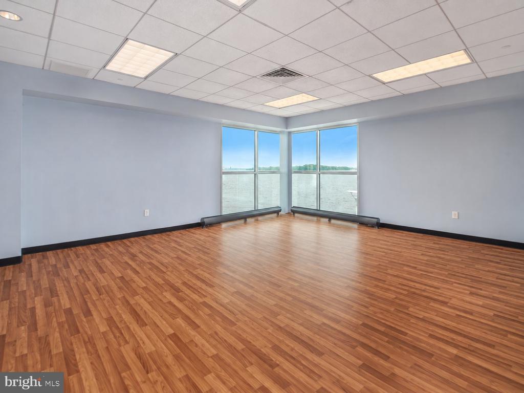 Empty corner room with wood flooring and tall windows overlooking water.