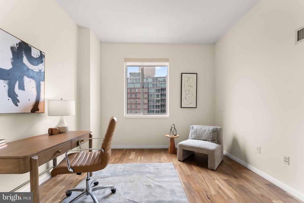 Furnished home office with a wooden desk, leather chair, and window overlooking nearby buildings.