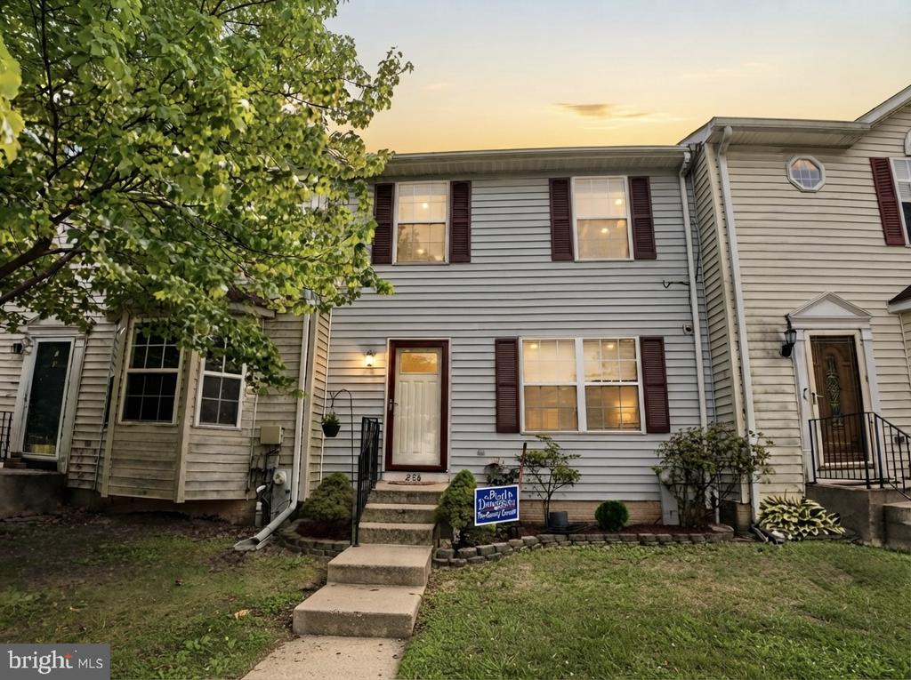 Two-story gray townhouse with burgundy shutters, front steps, and a large leafy tree.