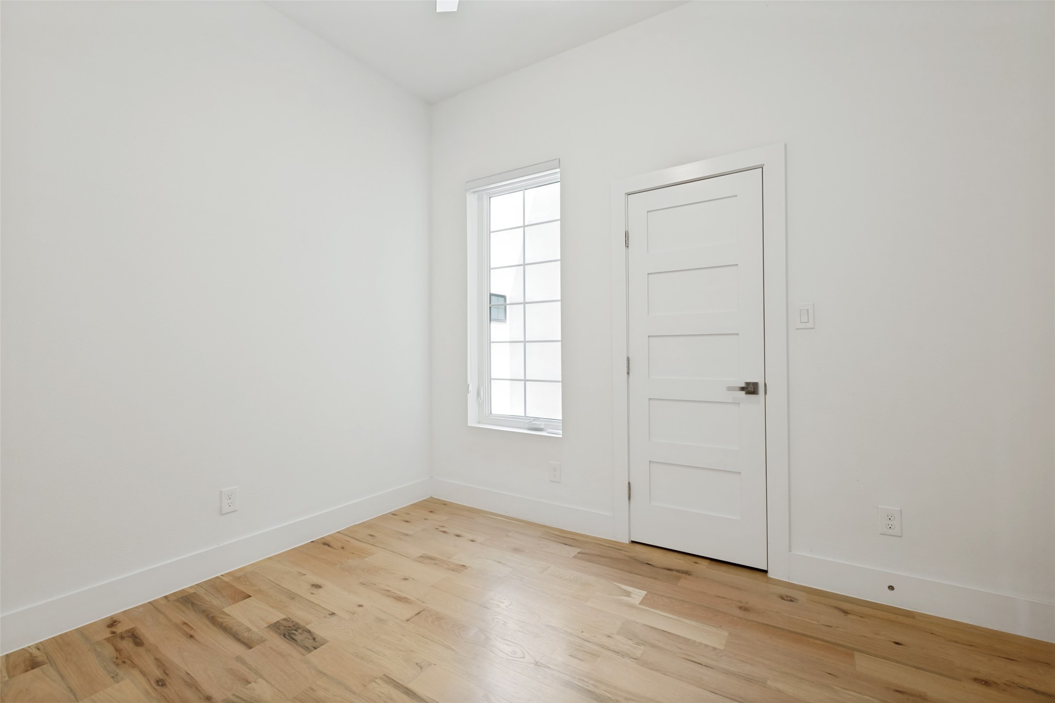 Empty room with natural wood floors, a tall divided-light window, and a white paneled door.