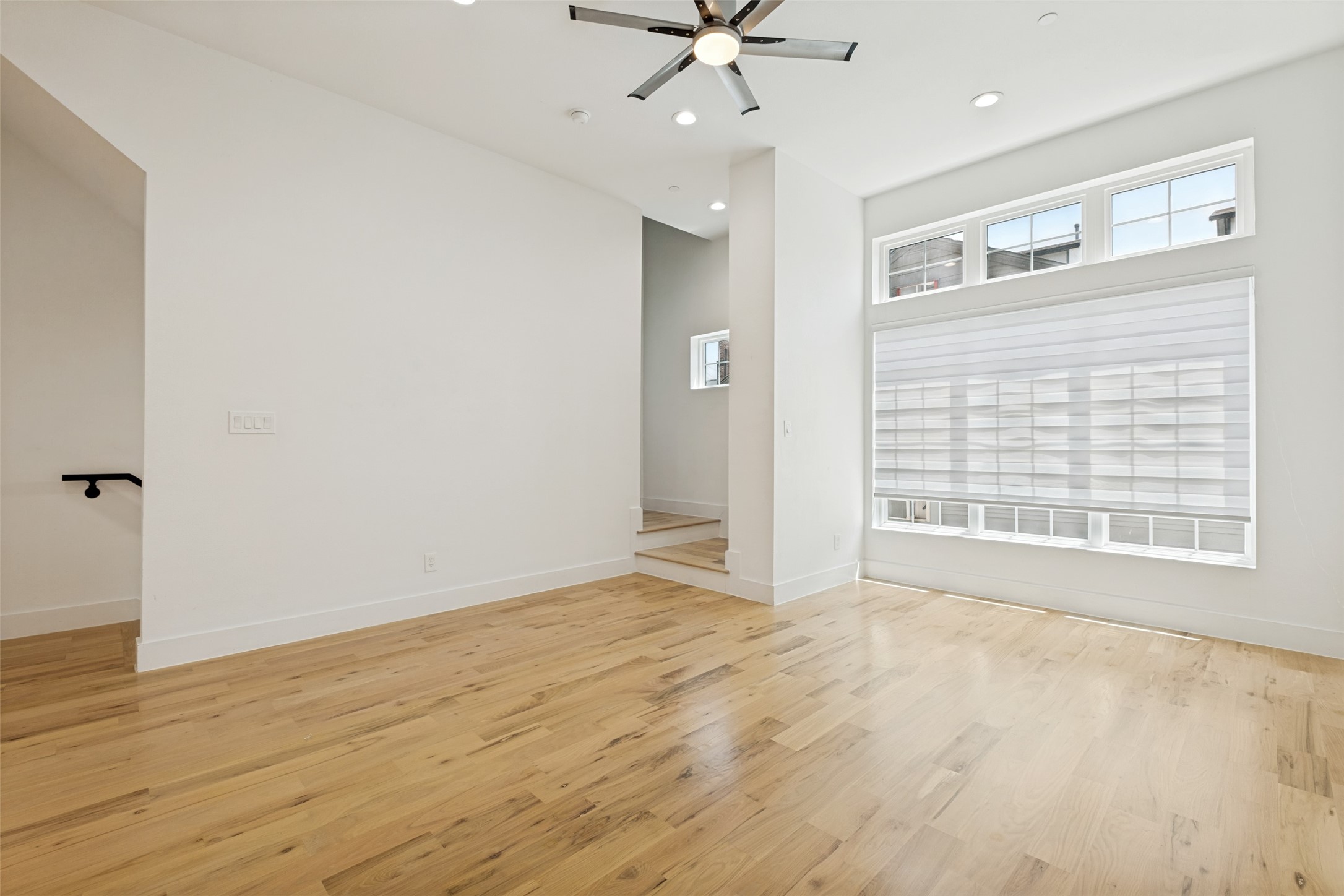 Empty living room with hardwood floors, large shaded window, and ceiling fan.