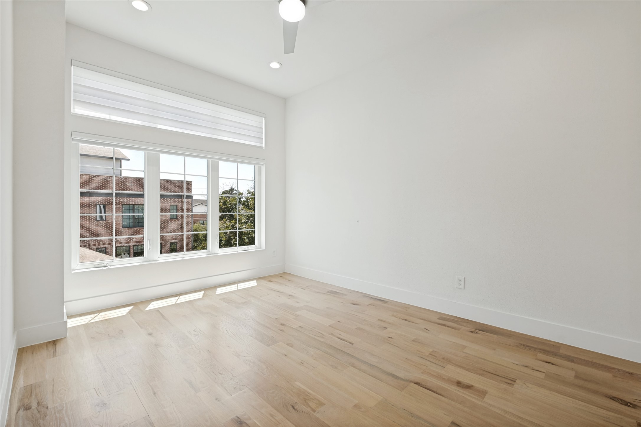 Empty room with pale wood floors and a large window overlooking a brick building.