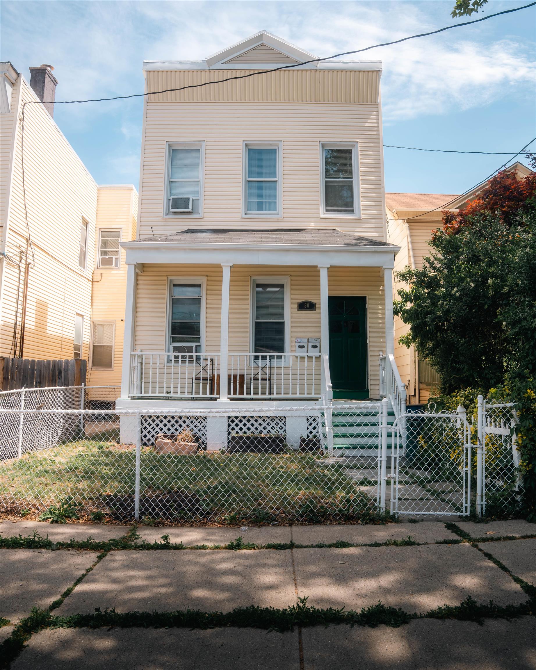 Light-colored multi-story house with a covered porch, dark green door, and fenced front yard.