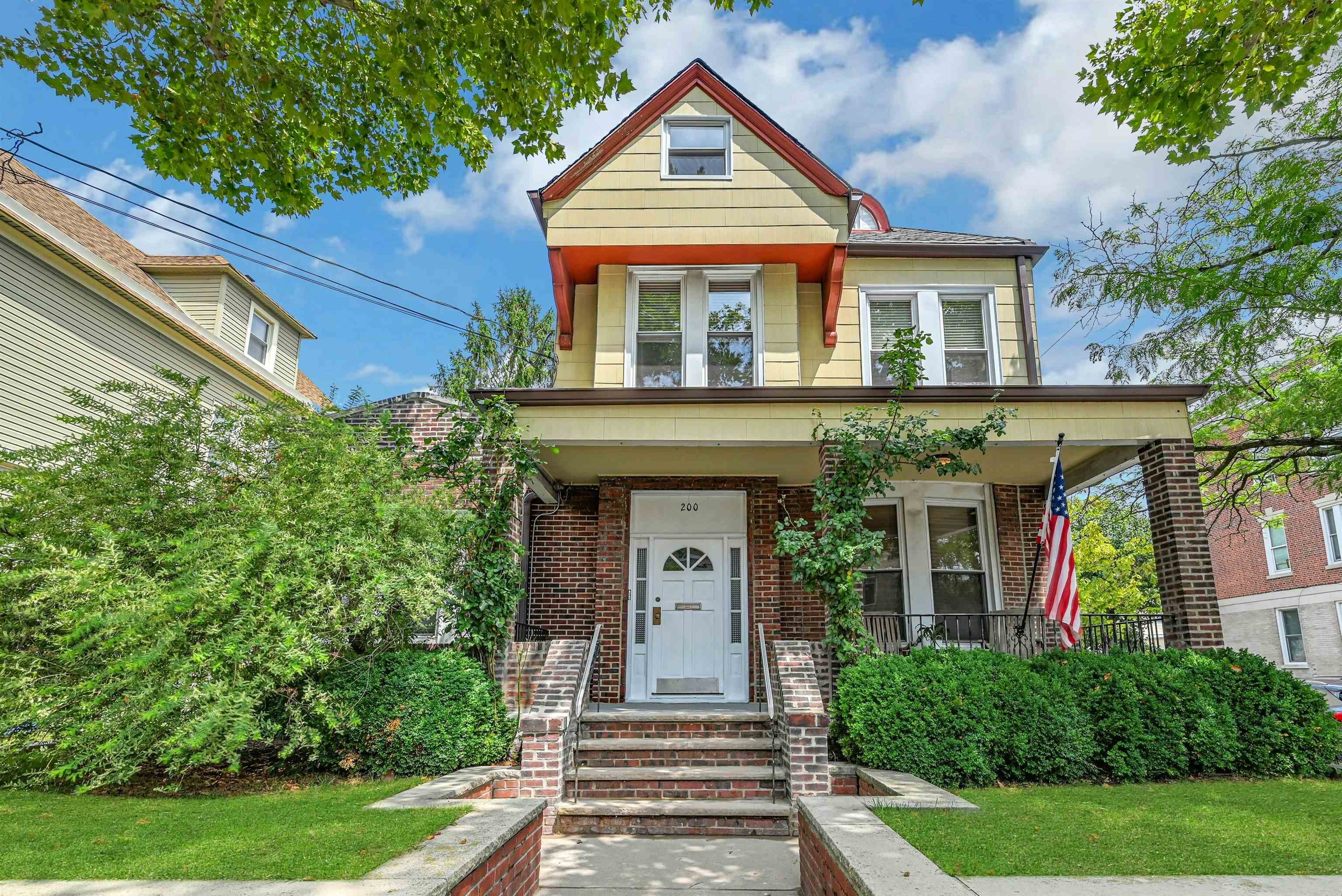 Yellow two-story house with red trim, brick porch, front steps, and an American flag.