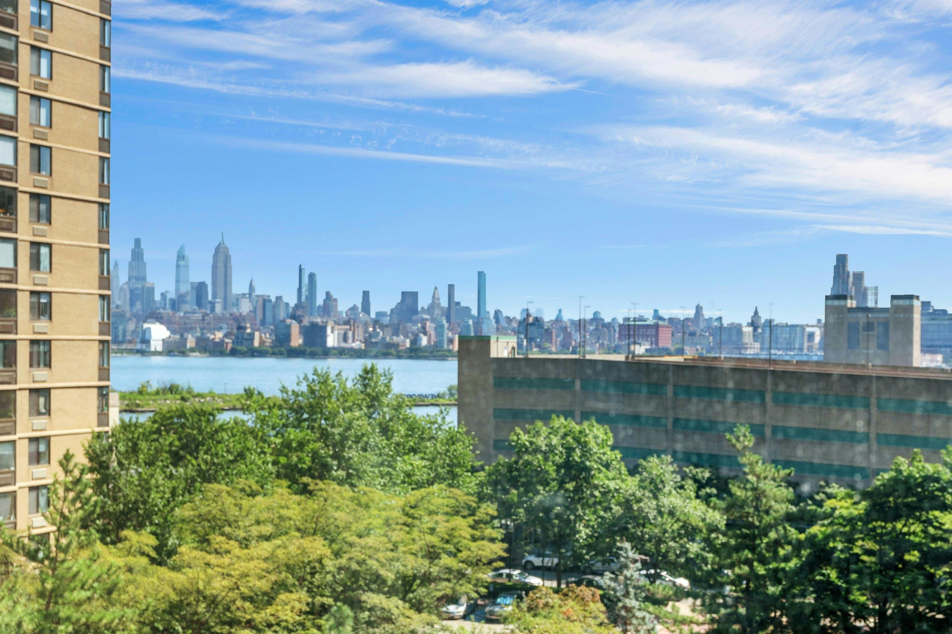 Manhattan skyline across the Hudson River, seen from Hoboken above leafy trees and nearby buildings.
