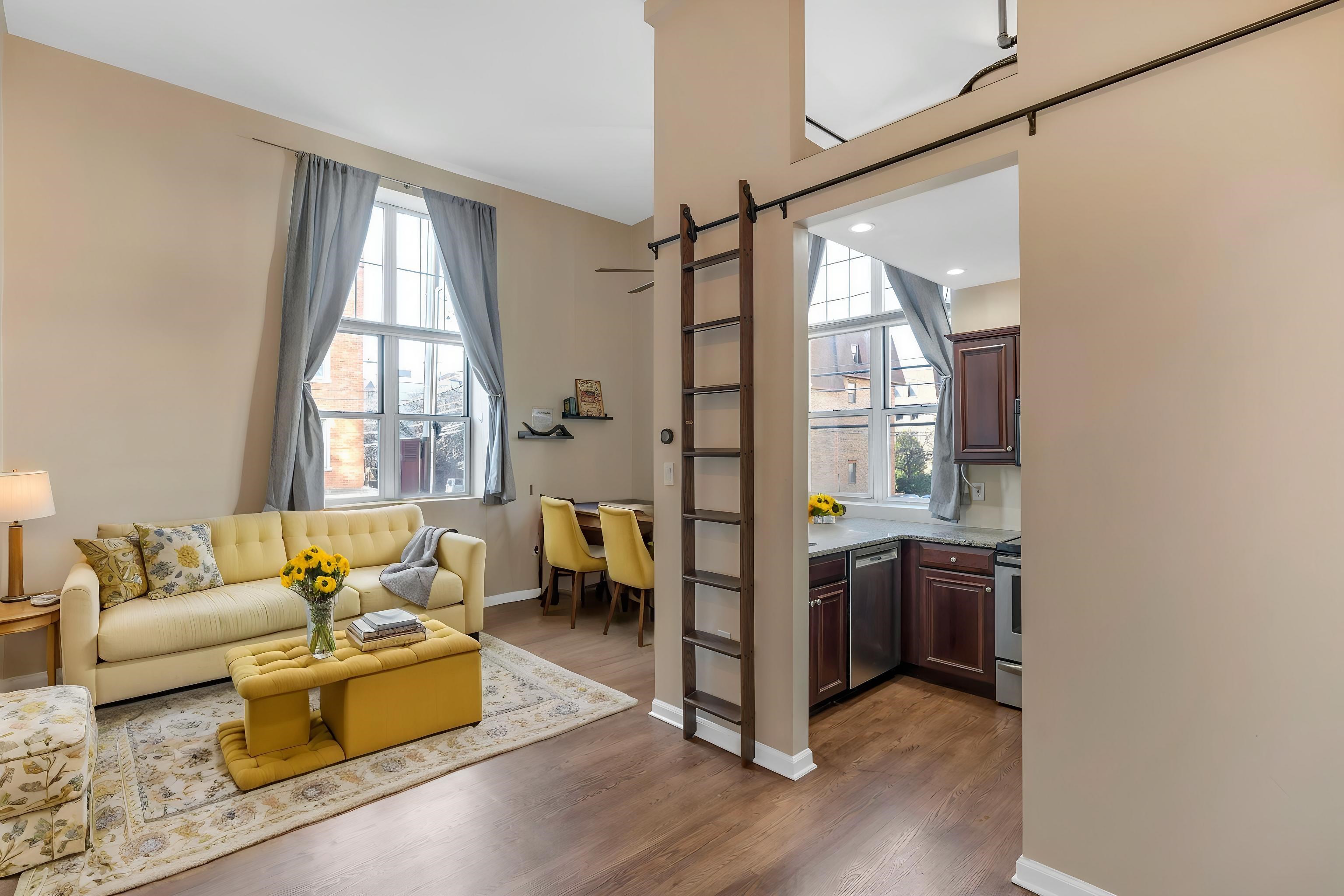 Living room with a yellow sofa and ottoman, tall curtained windows, and a ladder to a loft.