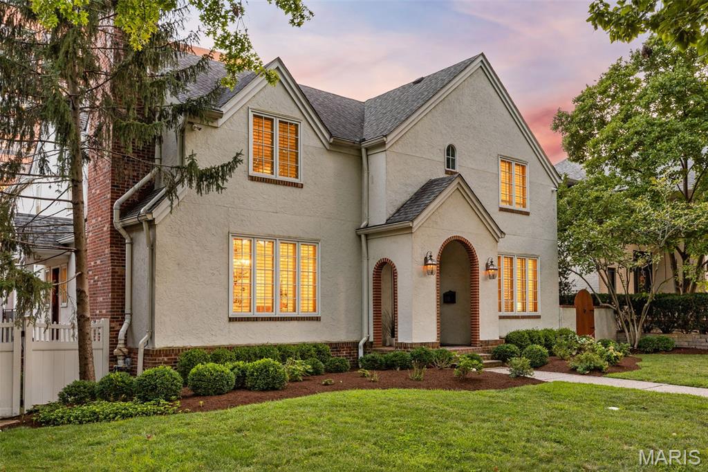 Two-story stucco home with an arched brick entry and warmly lit windows at dusk.